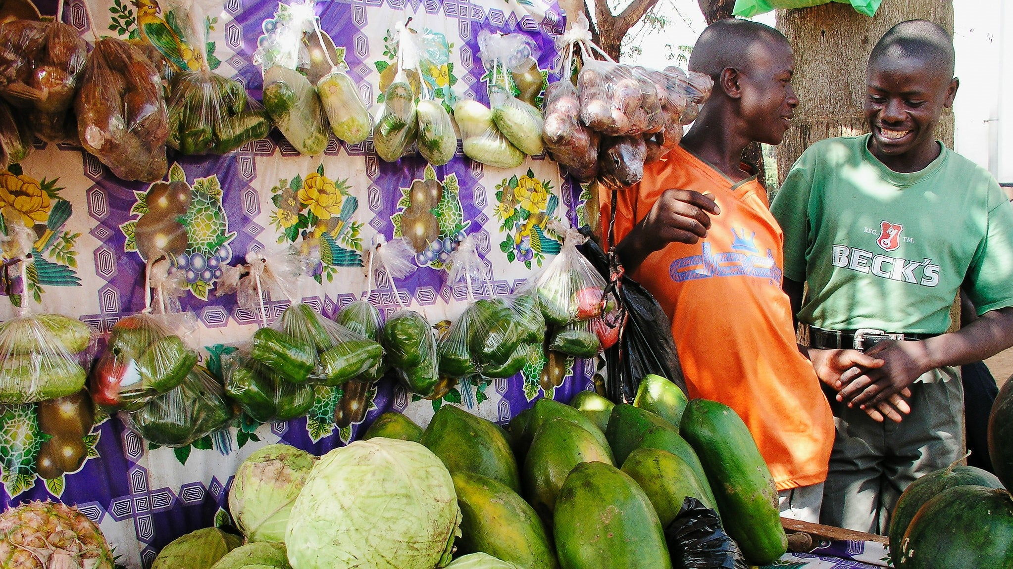 Two men smiling at a market stall with various fruits and vegetables.