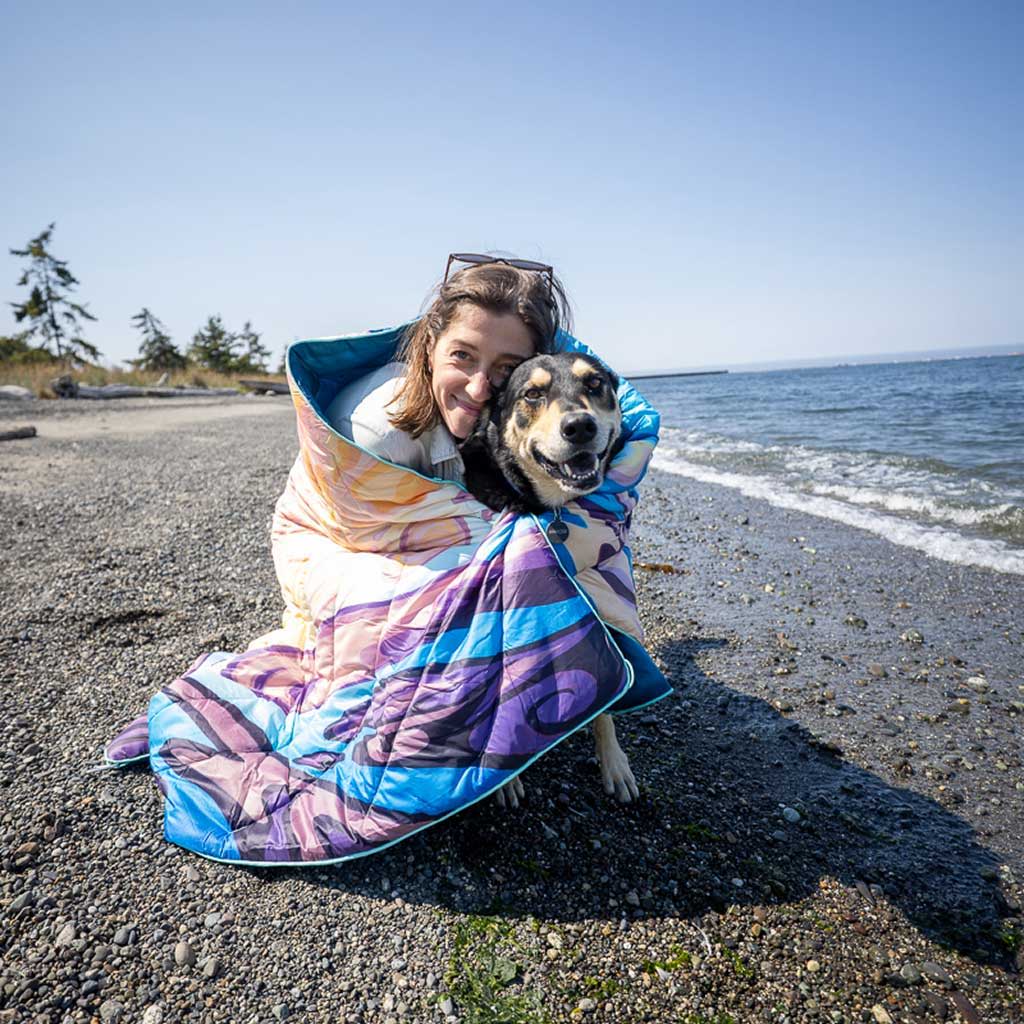 Gianna Andrews wrapped in a colorful blanket with a dog by the beach.