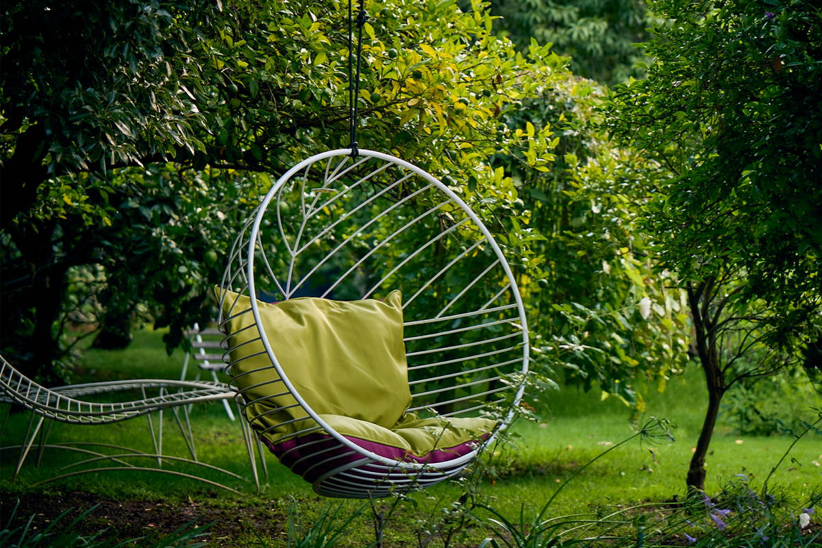 A bubble-shaped white wire chair filled with plush cushions hangs from a tree in a lush green garden.