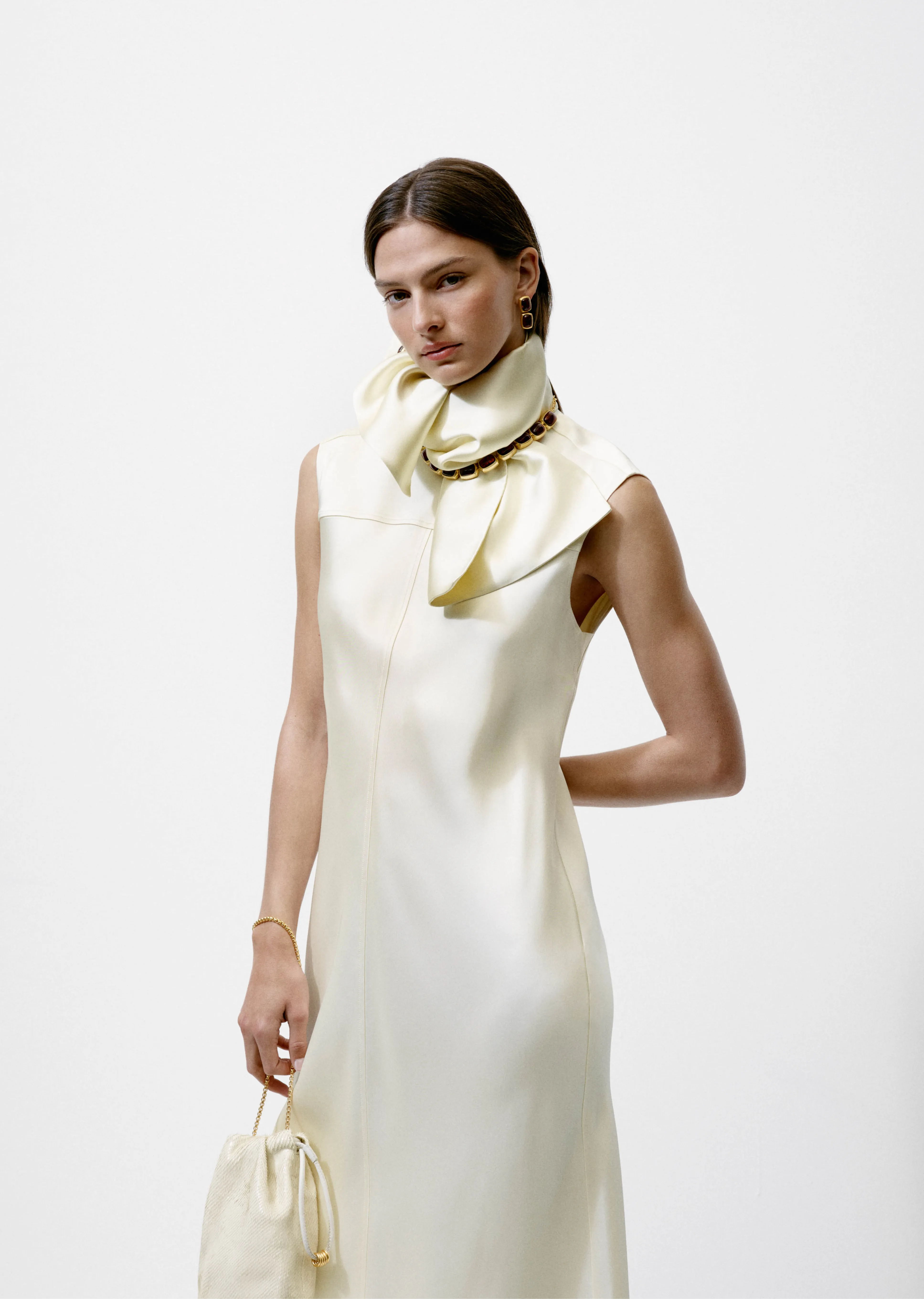 Model wearing a sleek, cream-colored dress with a matching scarf and handbag.