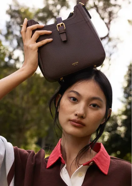 A person holding a brown handbag above their head outdoors.