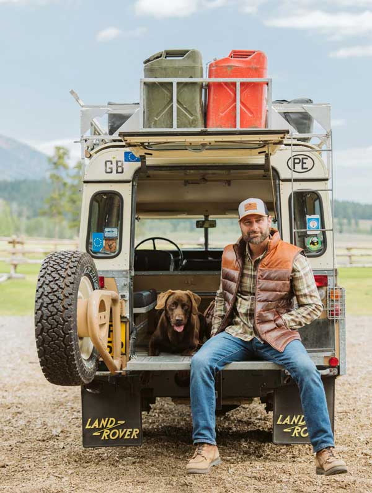 Man in a Buffalo Jackson leather down vest sitting on a vintage Land Rover tailgate with his chocolate Lab