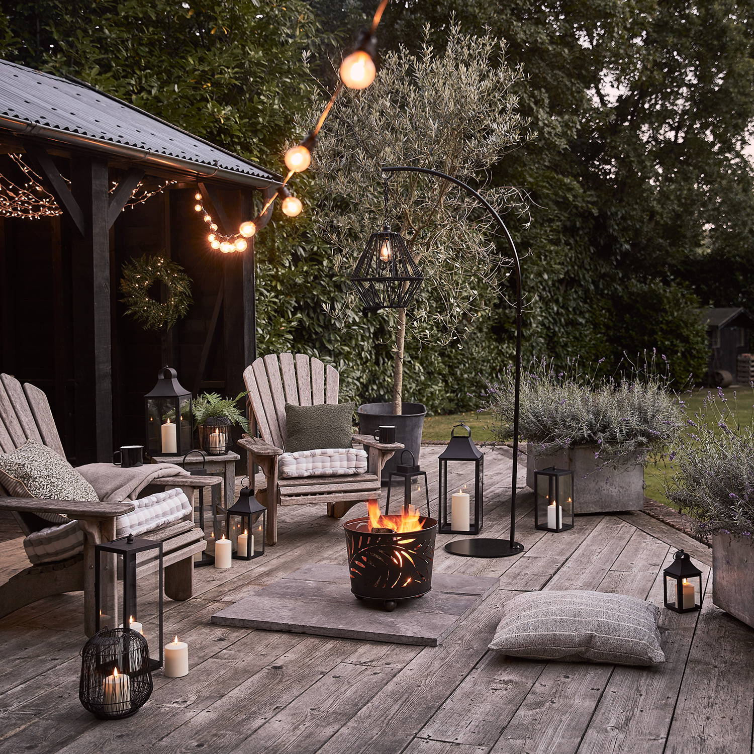 An outdoor summer seating area with LED candles and lanterns, a cosy fire bucket and warm white festoon lights hanging above.