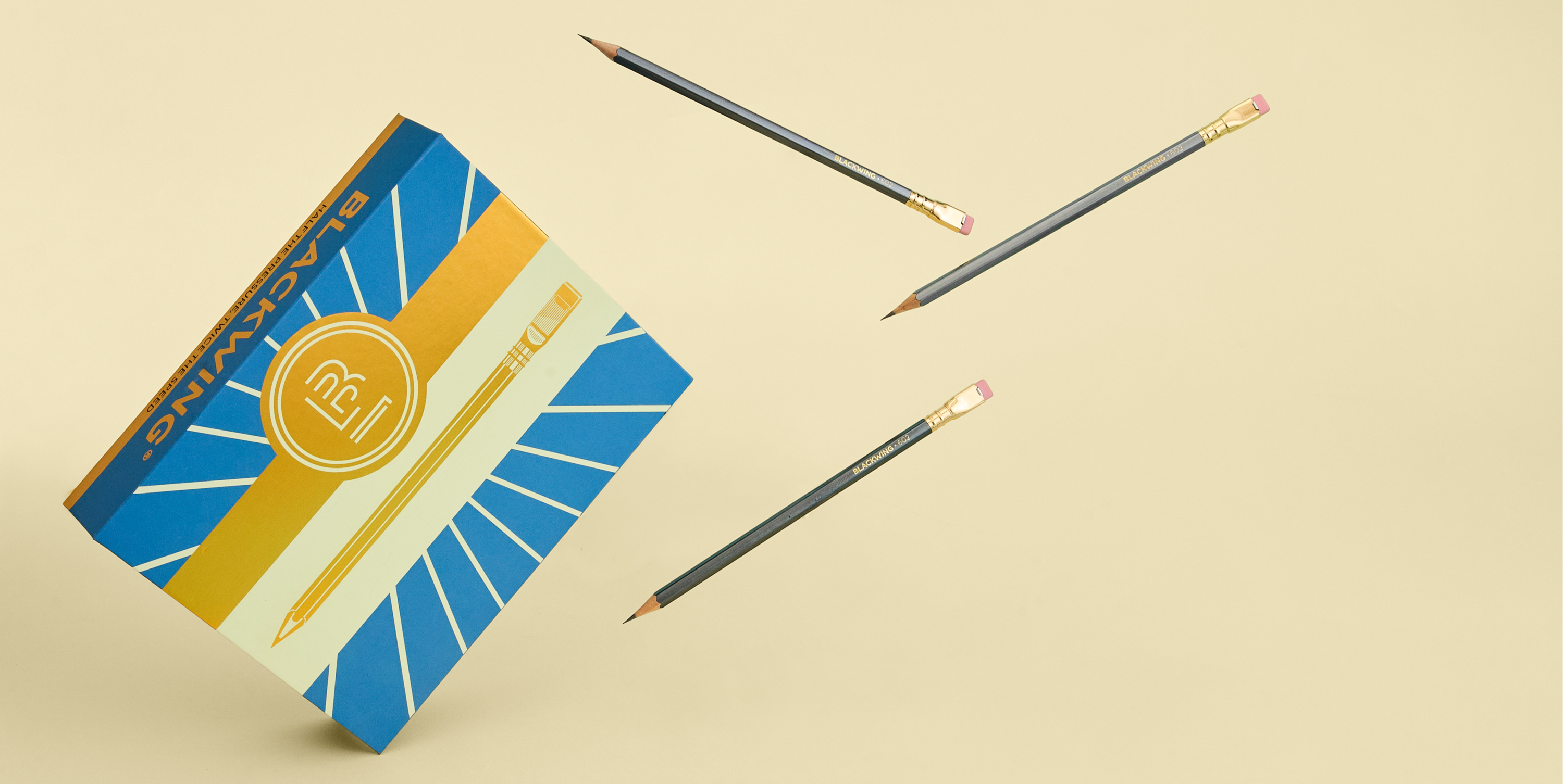 A blue gift box with a sun design and gold foil 'B' emblem and pencil next to three Blackwing 602 pencils on a beige background.