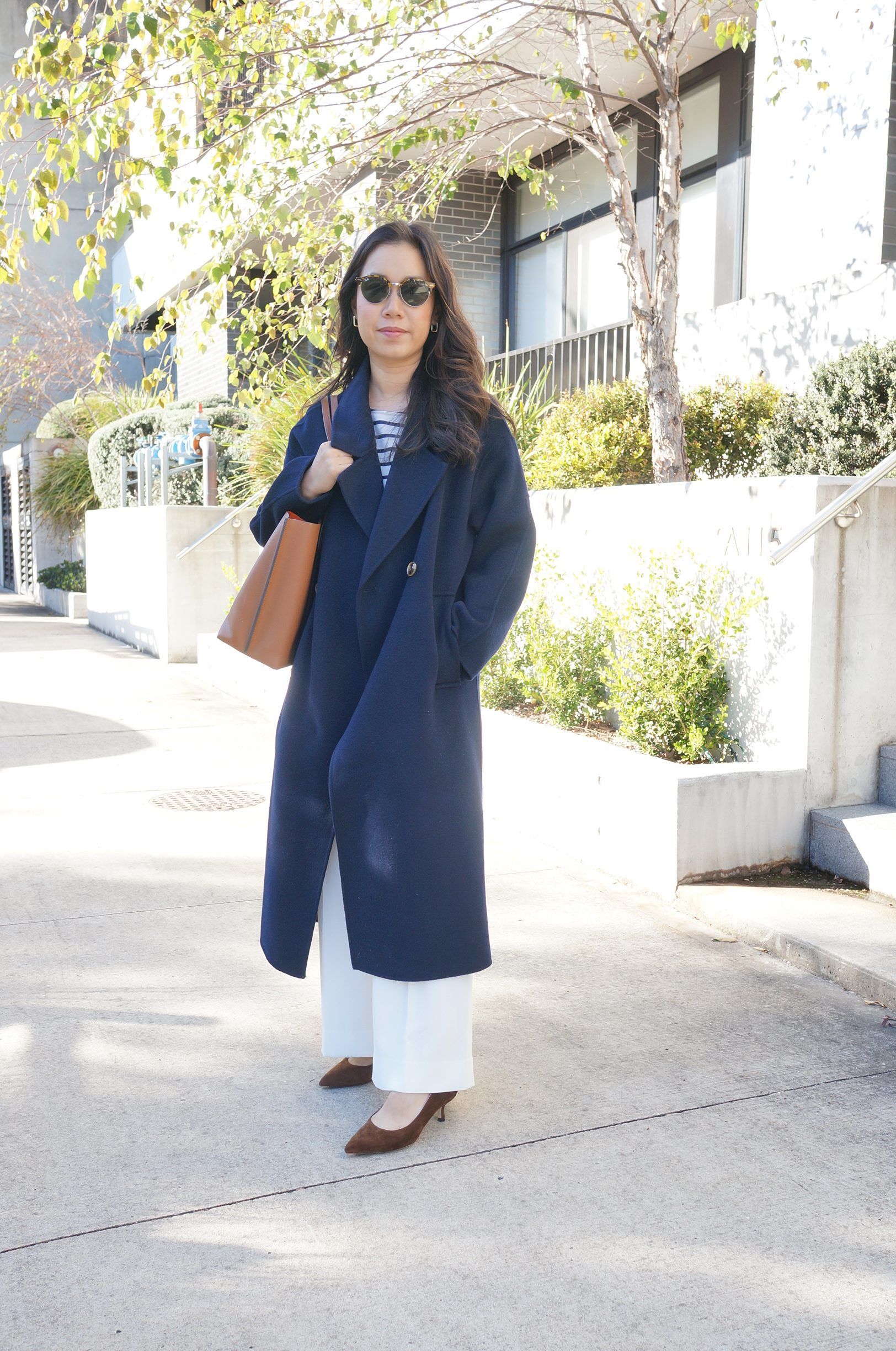 Woman in navy coat and sunglasses holding a brown bag.