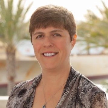 Smiling woman with short hair, wearing a patterned top, outdoors with palm trees.
