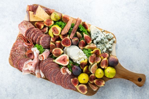 A wooden board with assorted meats, cheeses, fruits, and greens.