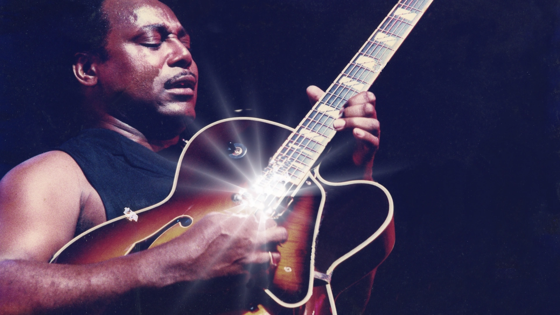 George Benson playing guitar with intense focus and light reflecting off the instrument.