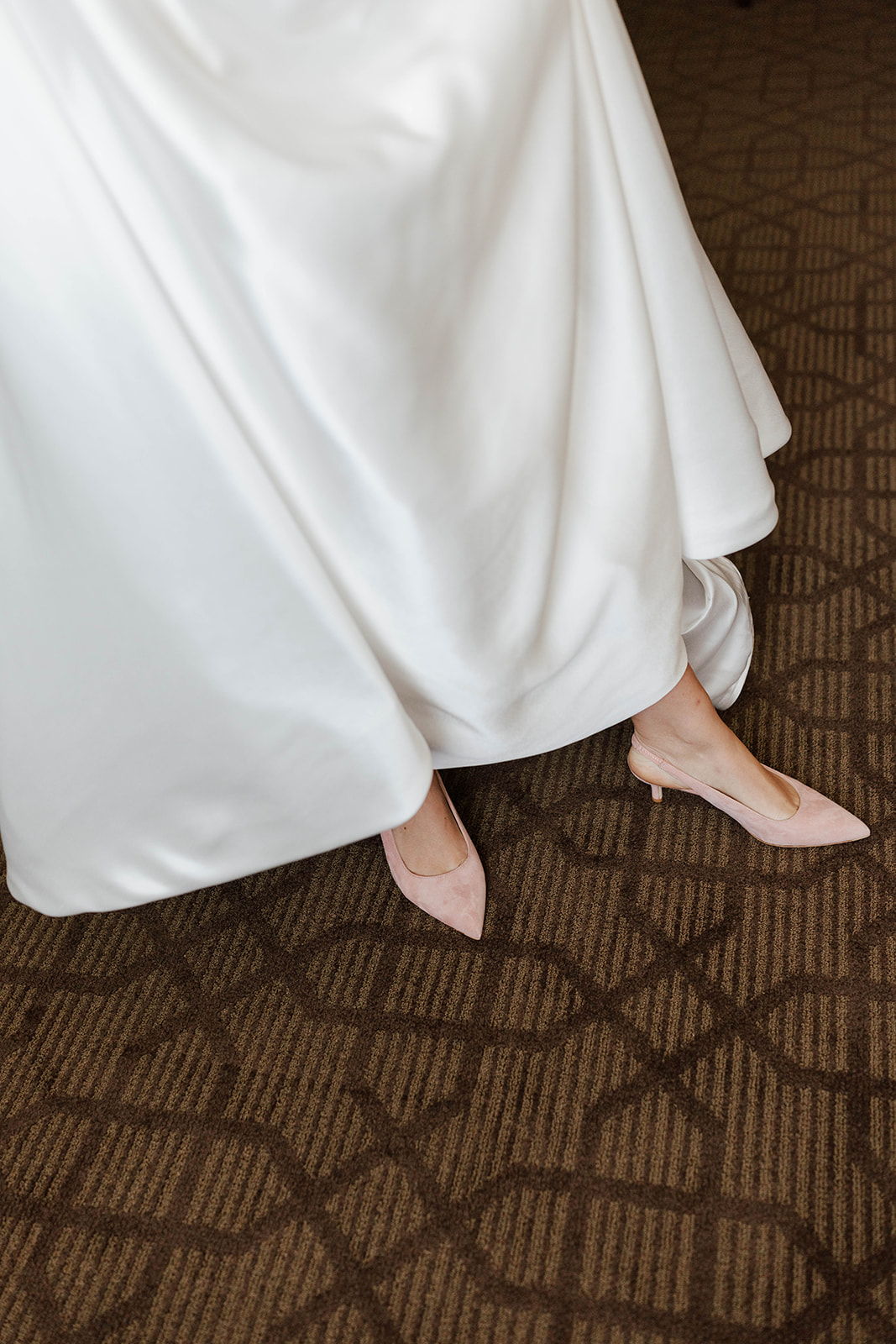 A close-up of a white gown and blush pink heels on a patterned carpet.
