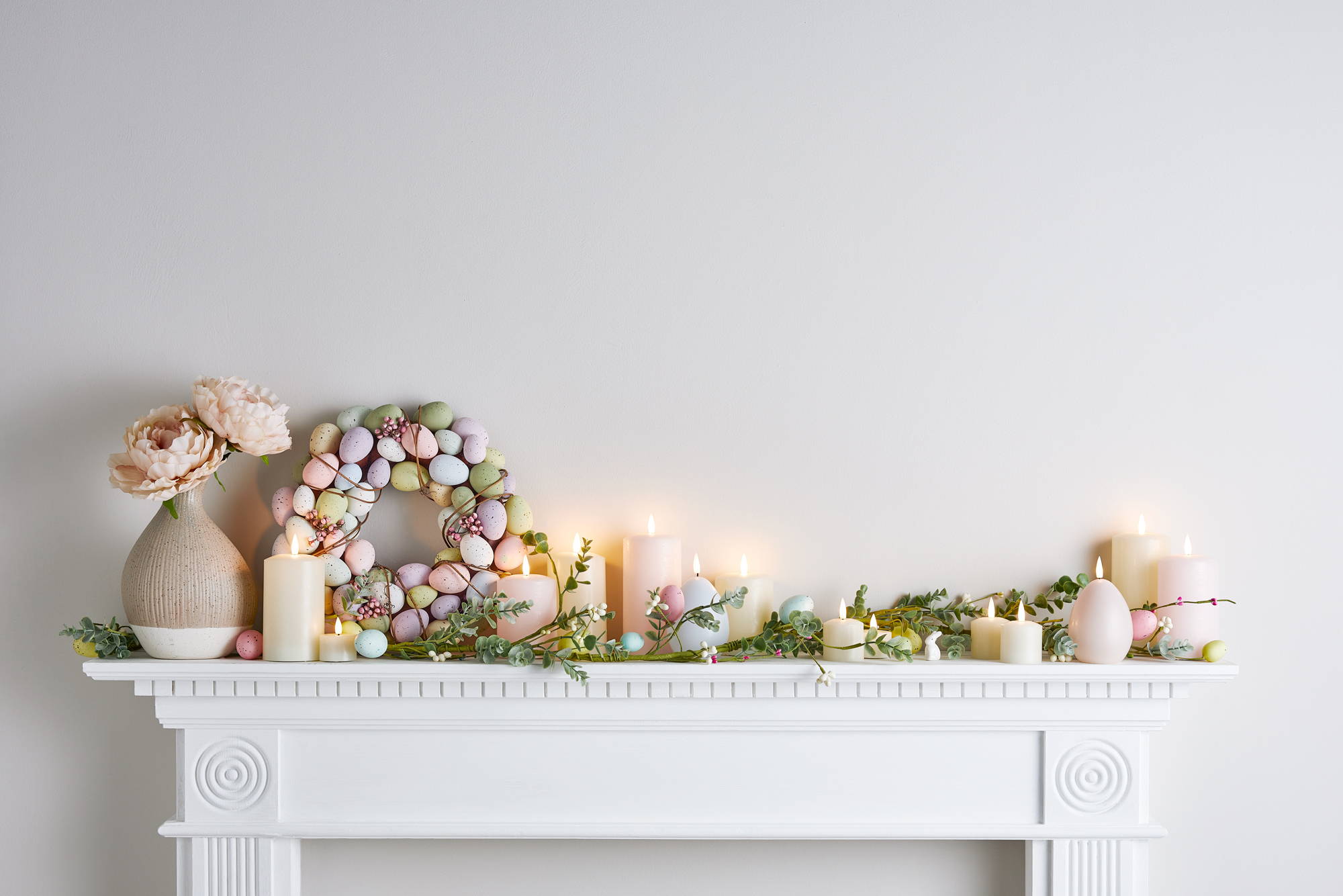 A white mantlepiece decorated with Easter wreaths, garland and pastel candles.