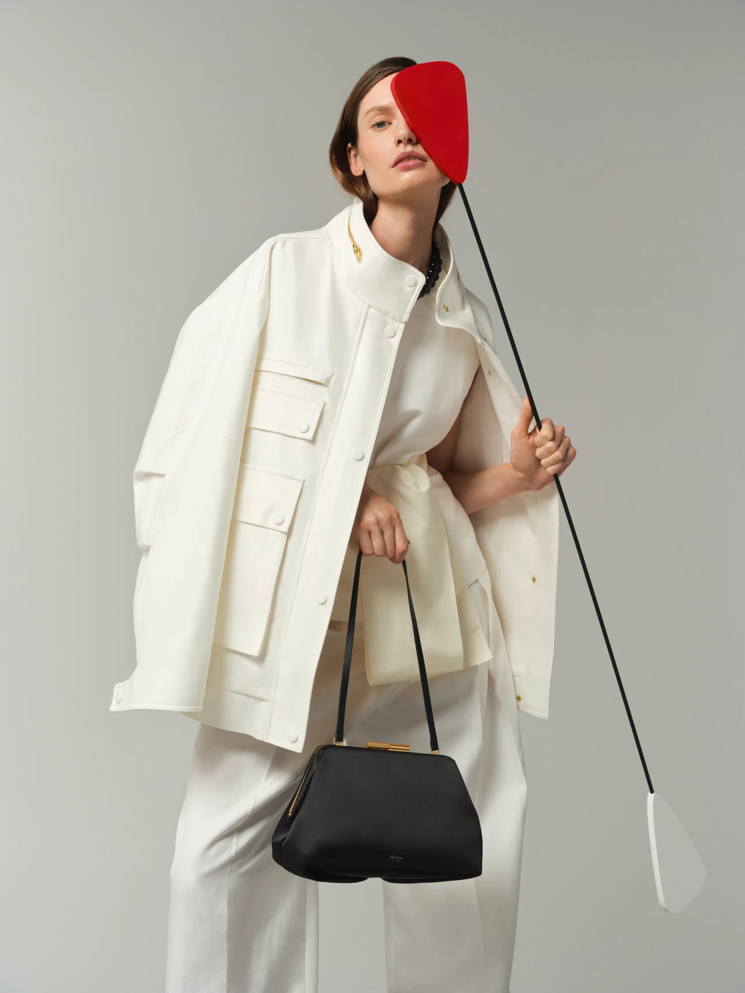 Model in white outfit holding a black bag and a red object.