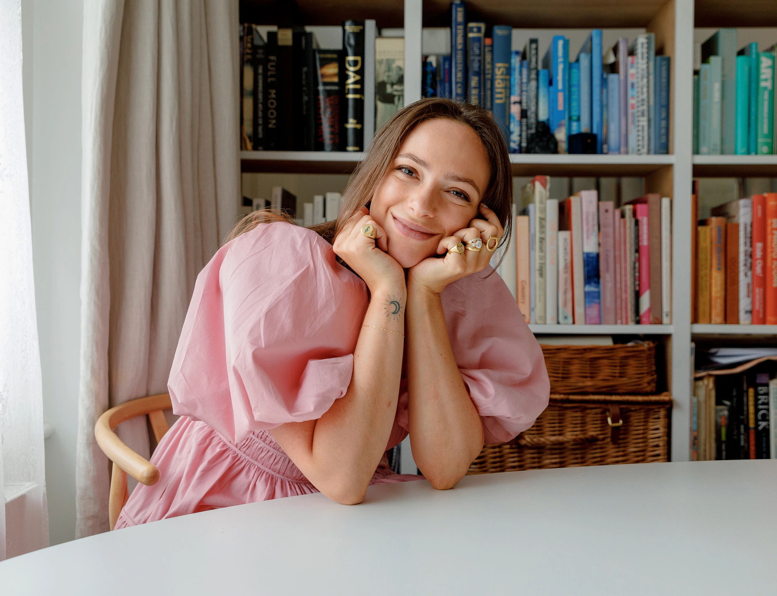Woman in pink dress smiling at a table with bookshelves in background.