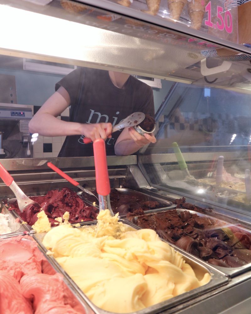 Person serving gelato from a display case with various flavors.