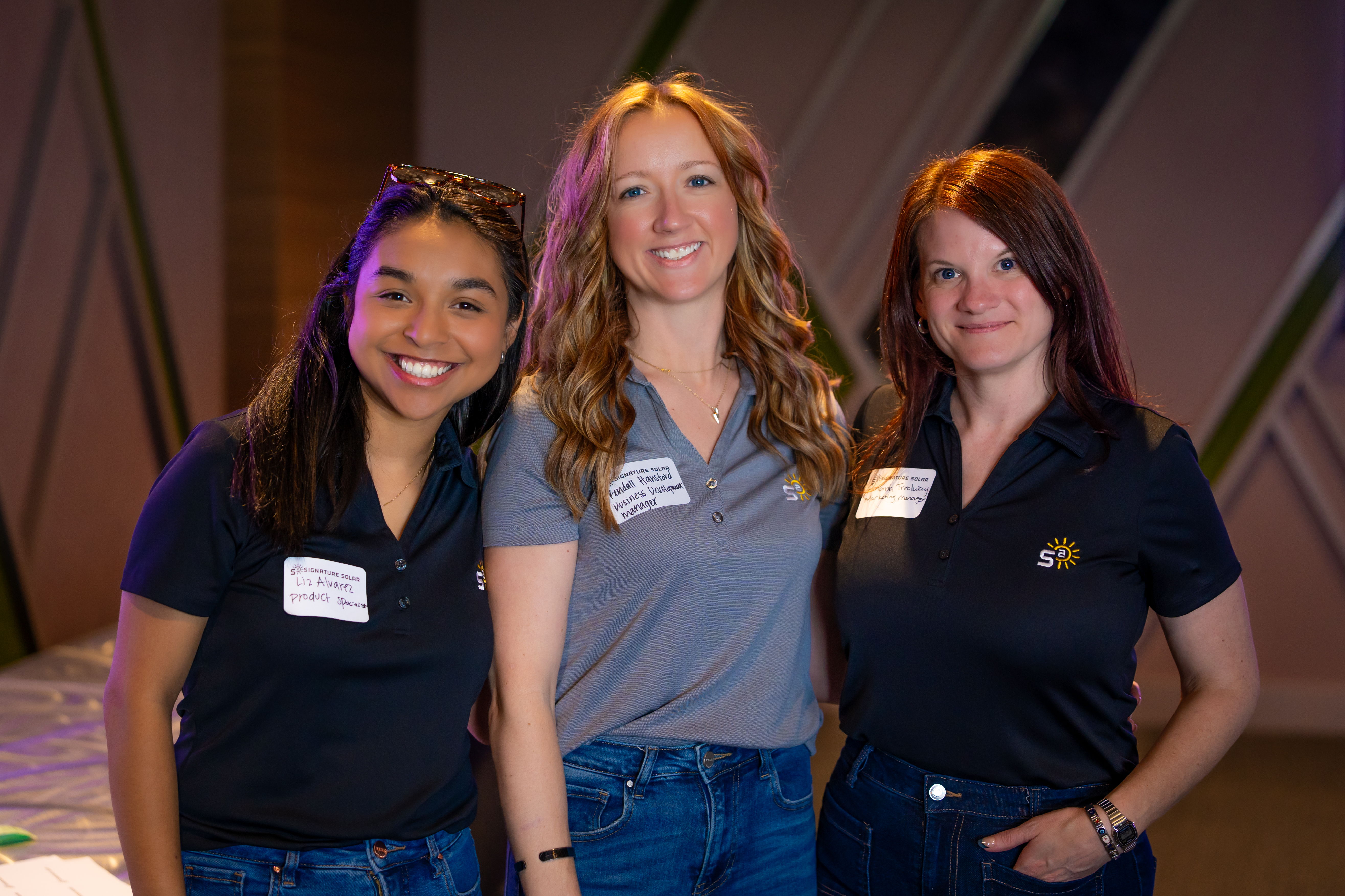Three women smiling together at an event, wearing matching polo shirts.