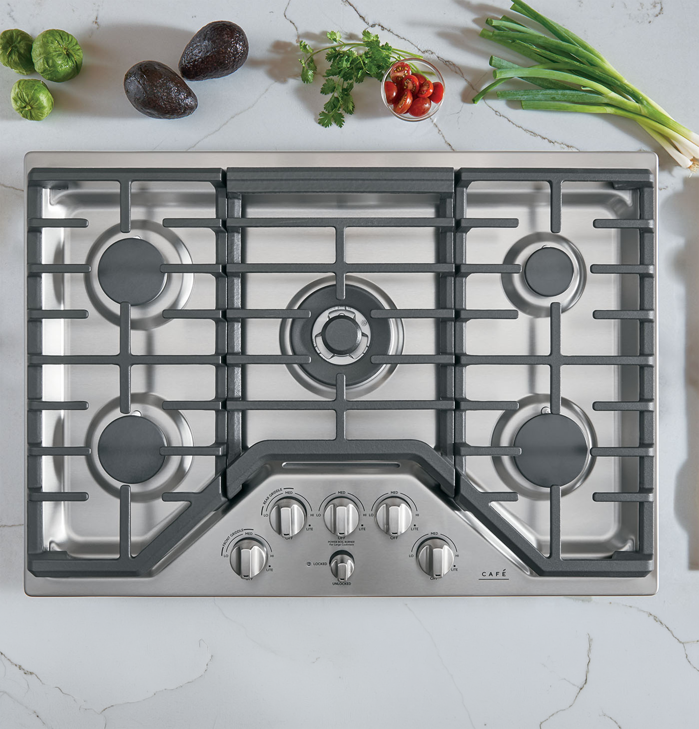 Stainless steel gas cooktop with knobs and surrounding vegetables on a countertop.