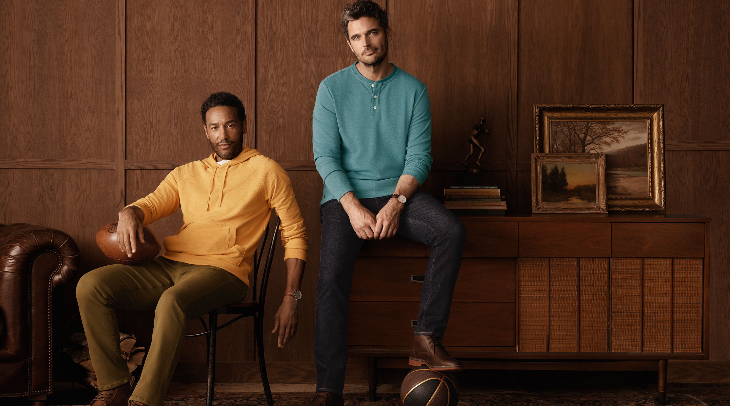 Two men seated, one in orange sweater, another in teal, against wooden backdrop.