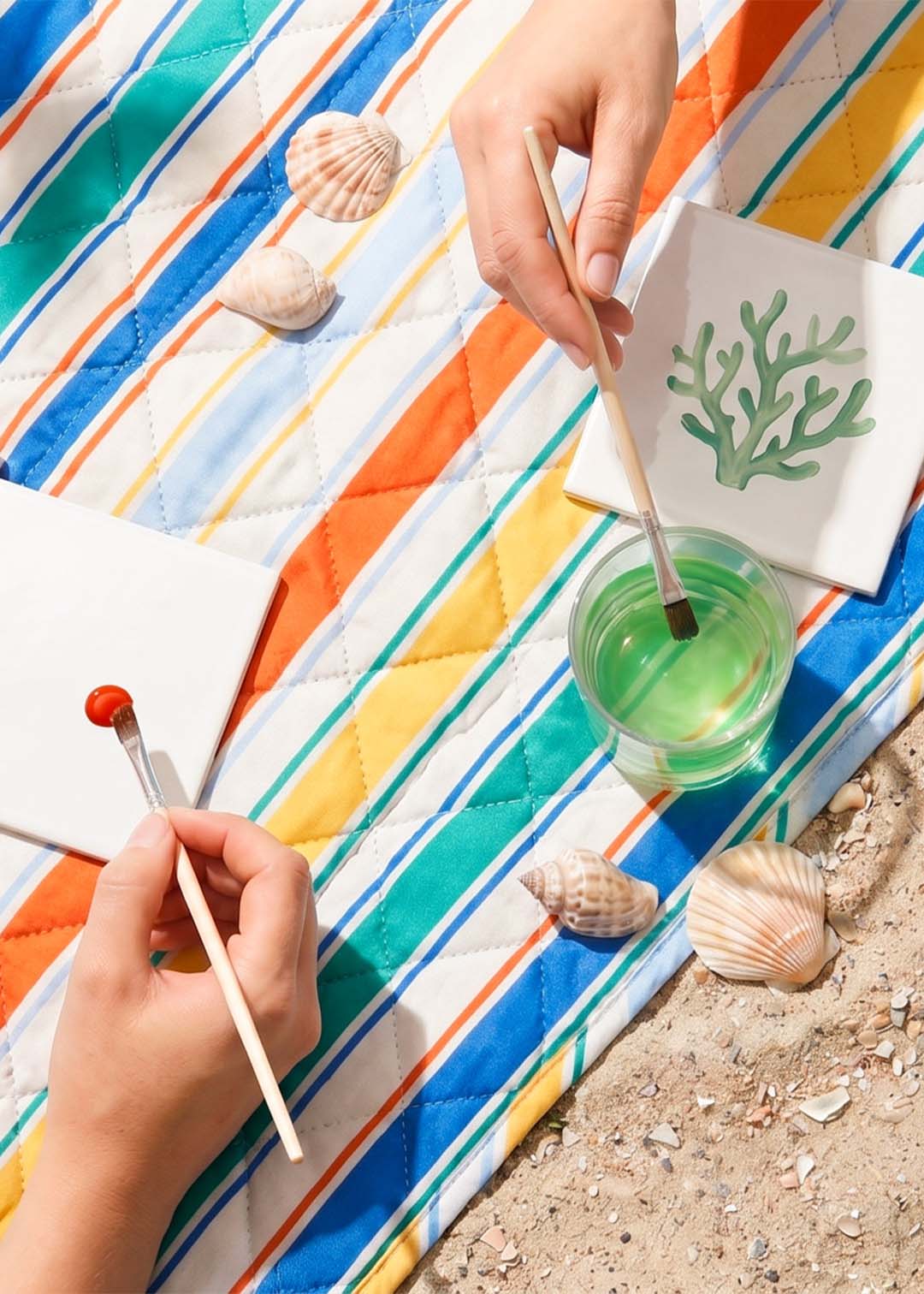 Hands painting on tiles with seashells and a paintbrush on a striped blanket.