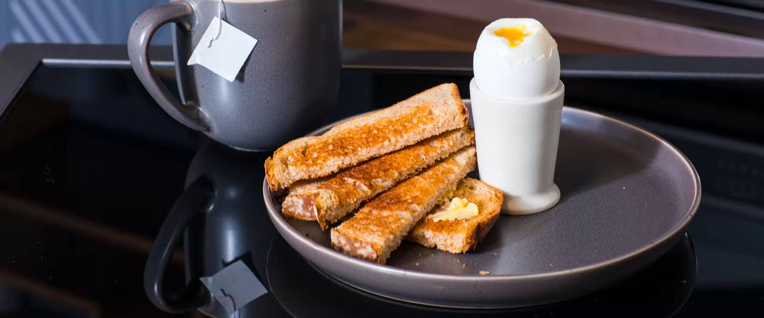 Soft boiled egg in an egg cup on a plate, with a side of sliced toast. Cup of tea in the background.