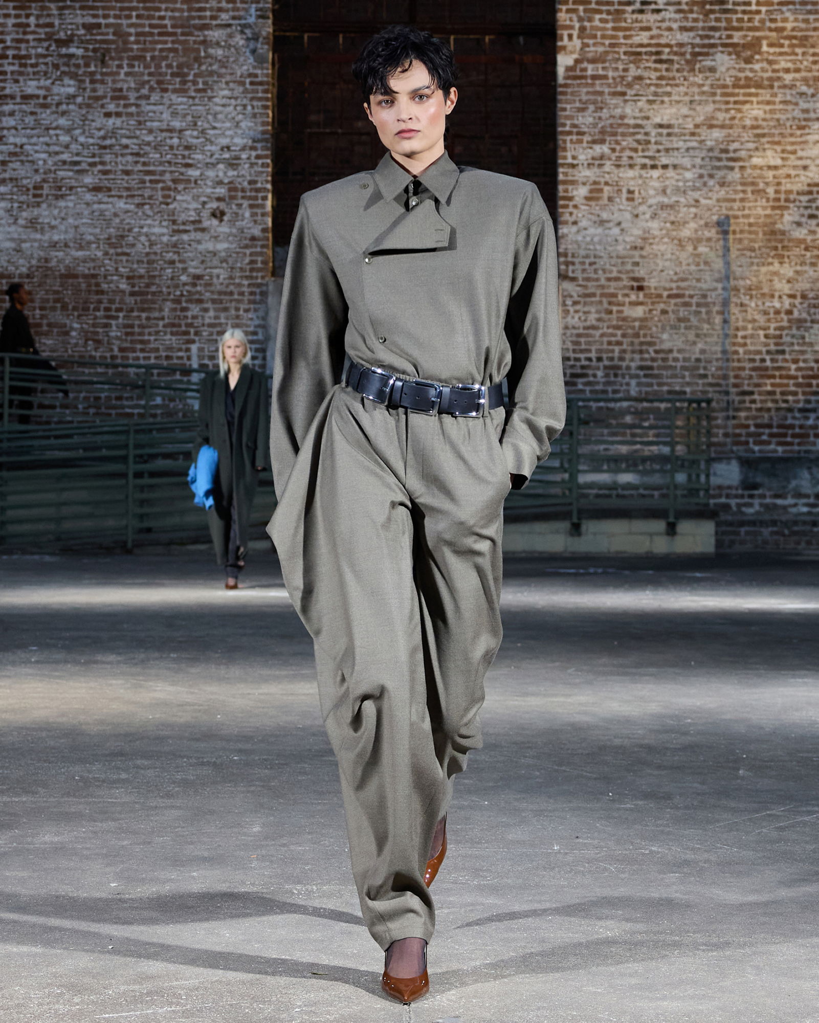 Model walking in oversized gray outfit with a belt in an industrial setting.