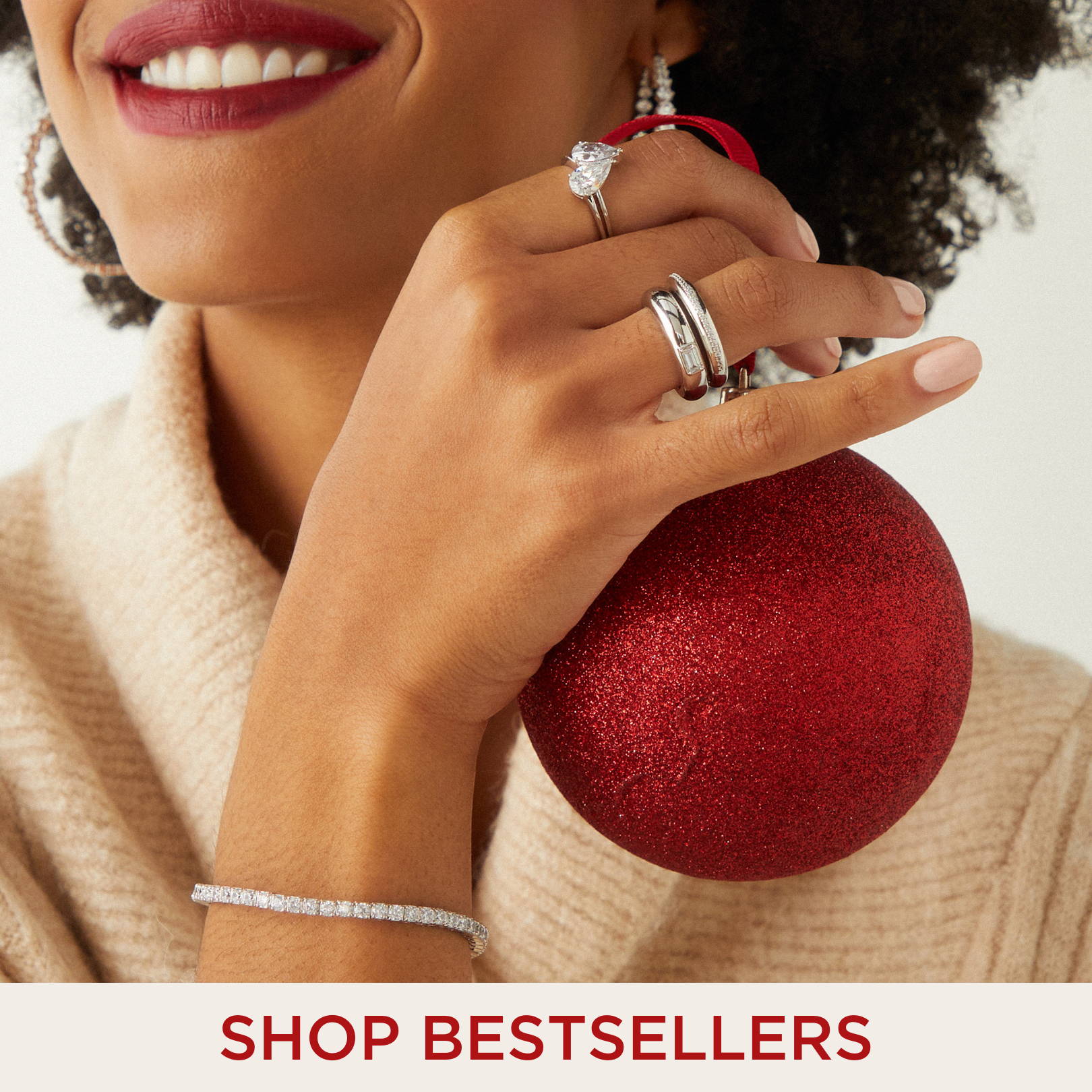 SHOP BESTSELLERS. Image of model wearing cubic zirconia rings and a cubic zirconia tennis bracelet while holding a red Christmas ornament. 