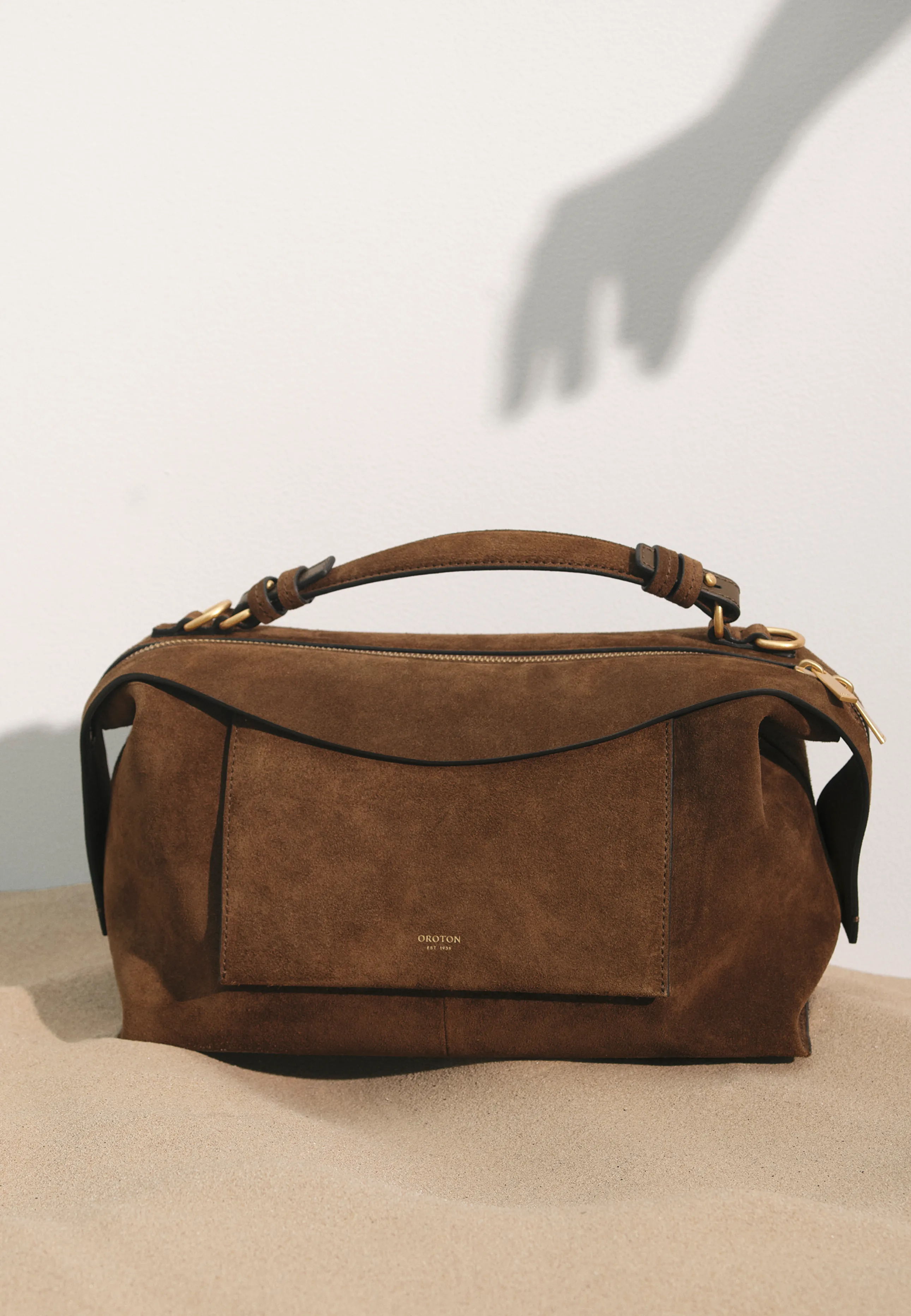 Brown suede handbag on a surface with a shadowed hand in the background.