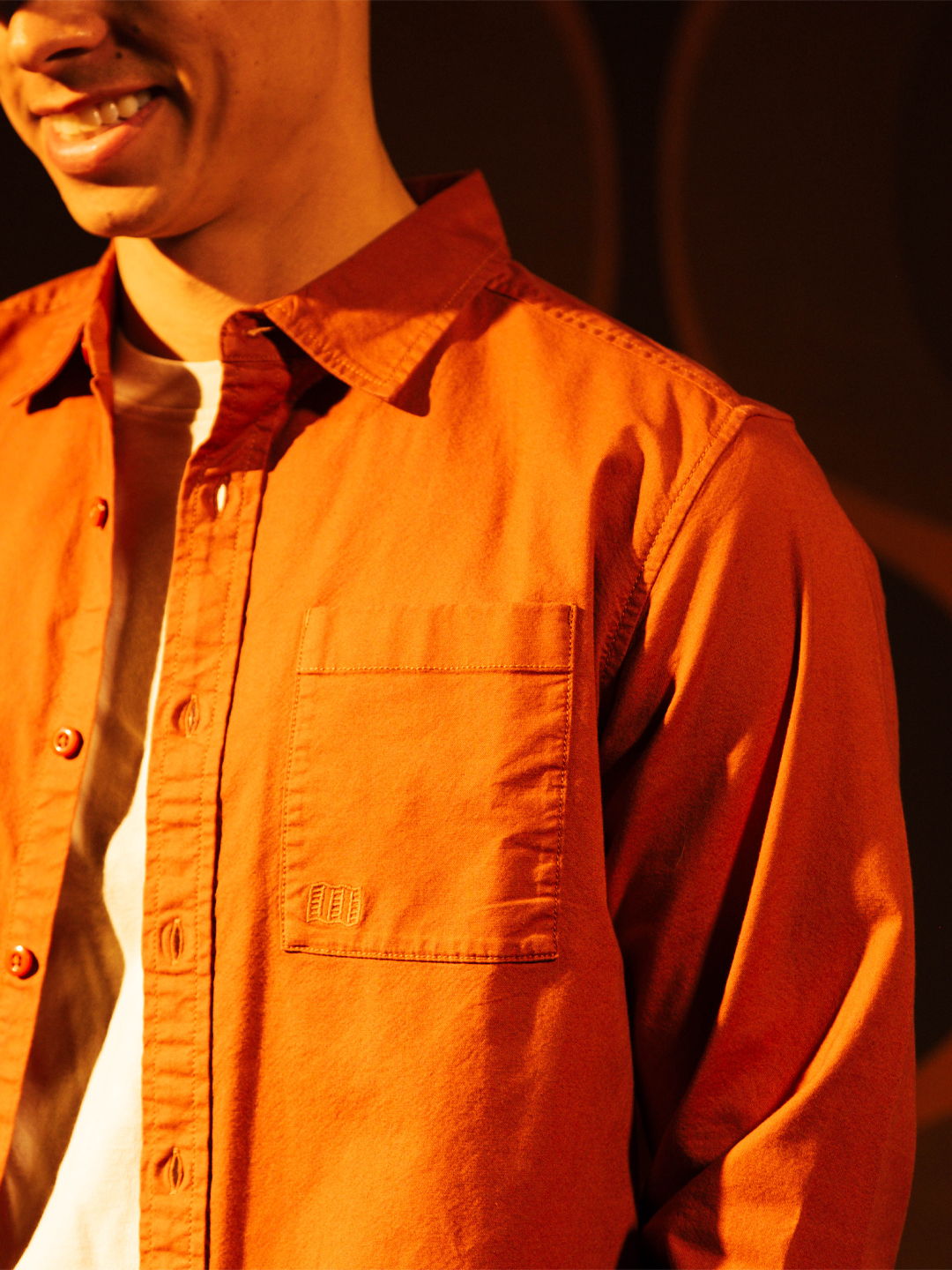 A person smiling while wearing an orange shirt and white t-shirt underneath.