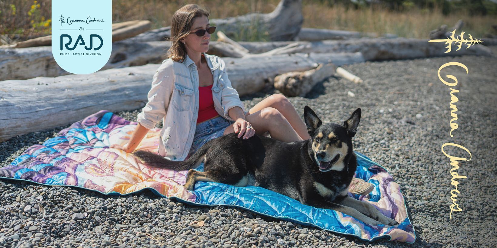 Woman sitting on a blanket with a dog on a pebbly beach.