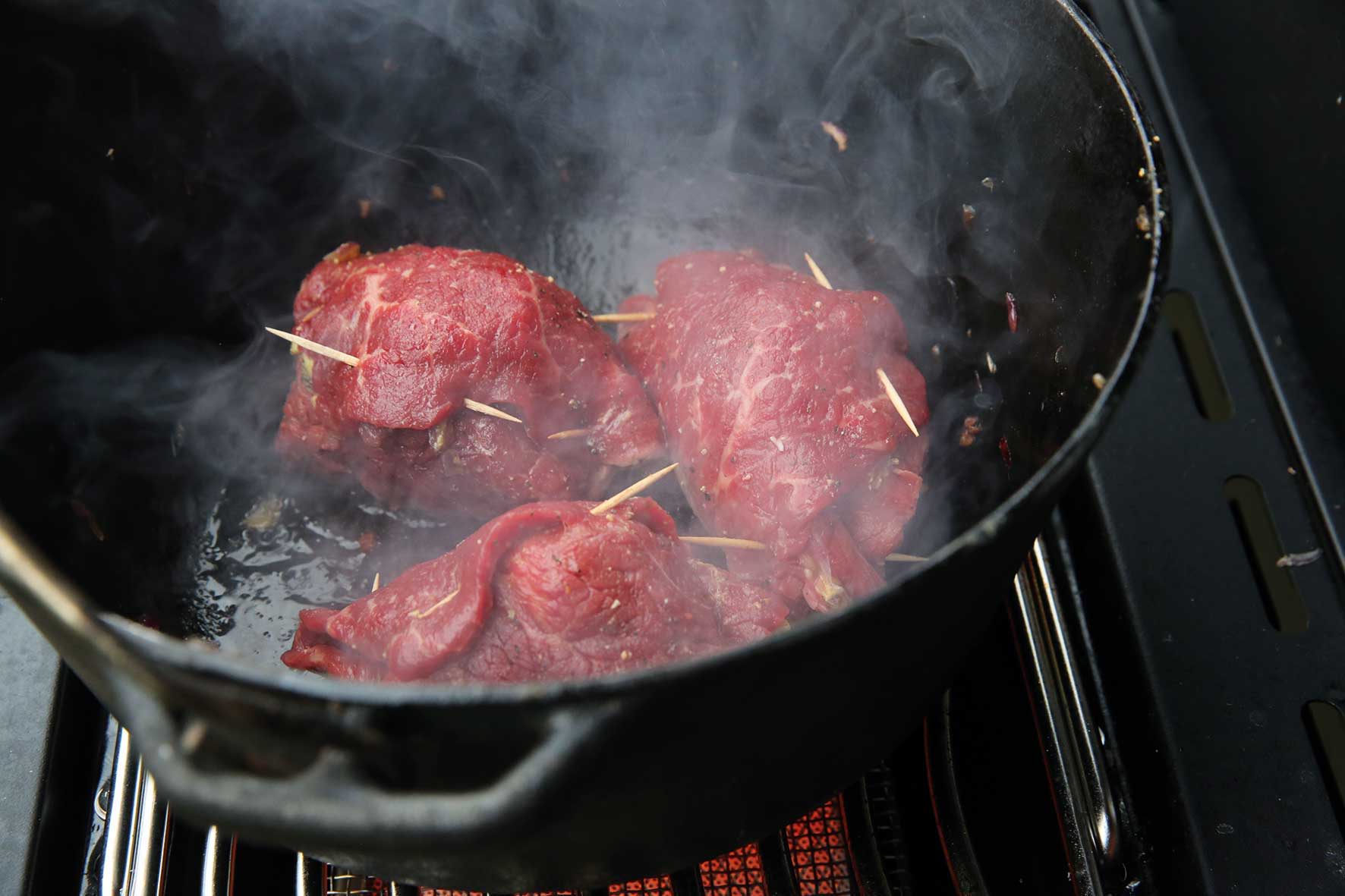 Raw meat skewered and cooking in a pot with smoke.
