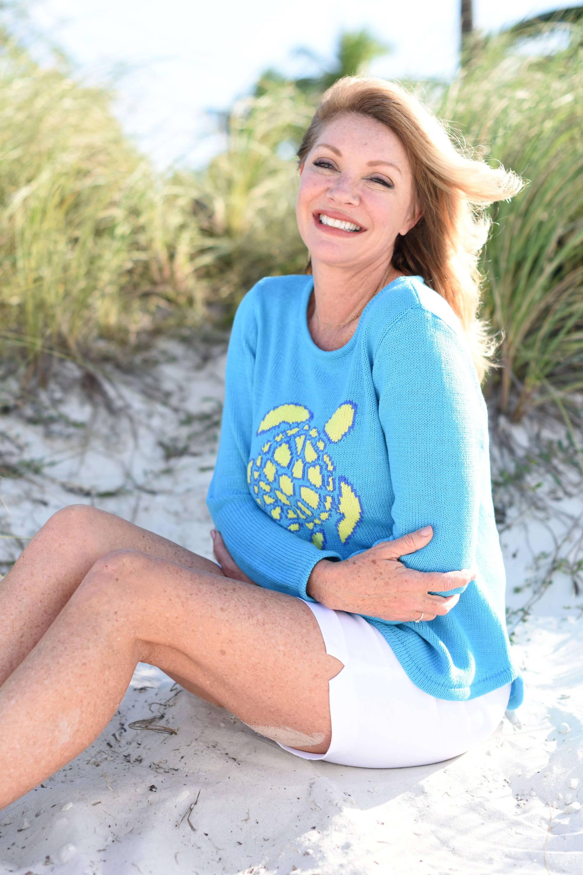 Smiling blonde woman sits on the sand as she hugs her arms to her chest. 