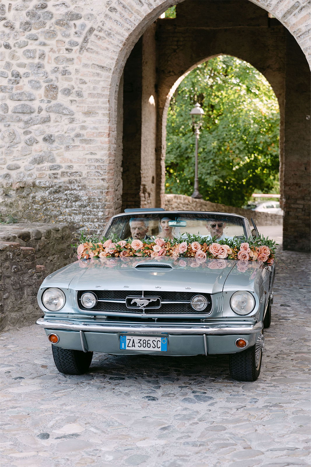 Vintage convertible car decorated with flowers, parked under a stone archway.
