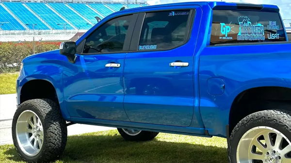 Side view of Carlos's blue 2022 Chevrolet Silverado 1500 with large polished wheels and sponsor decals near a stadium, install photo 1