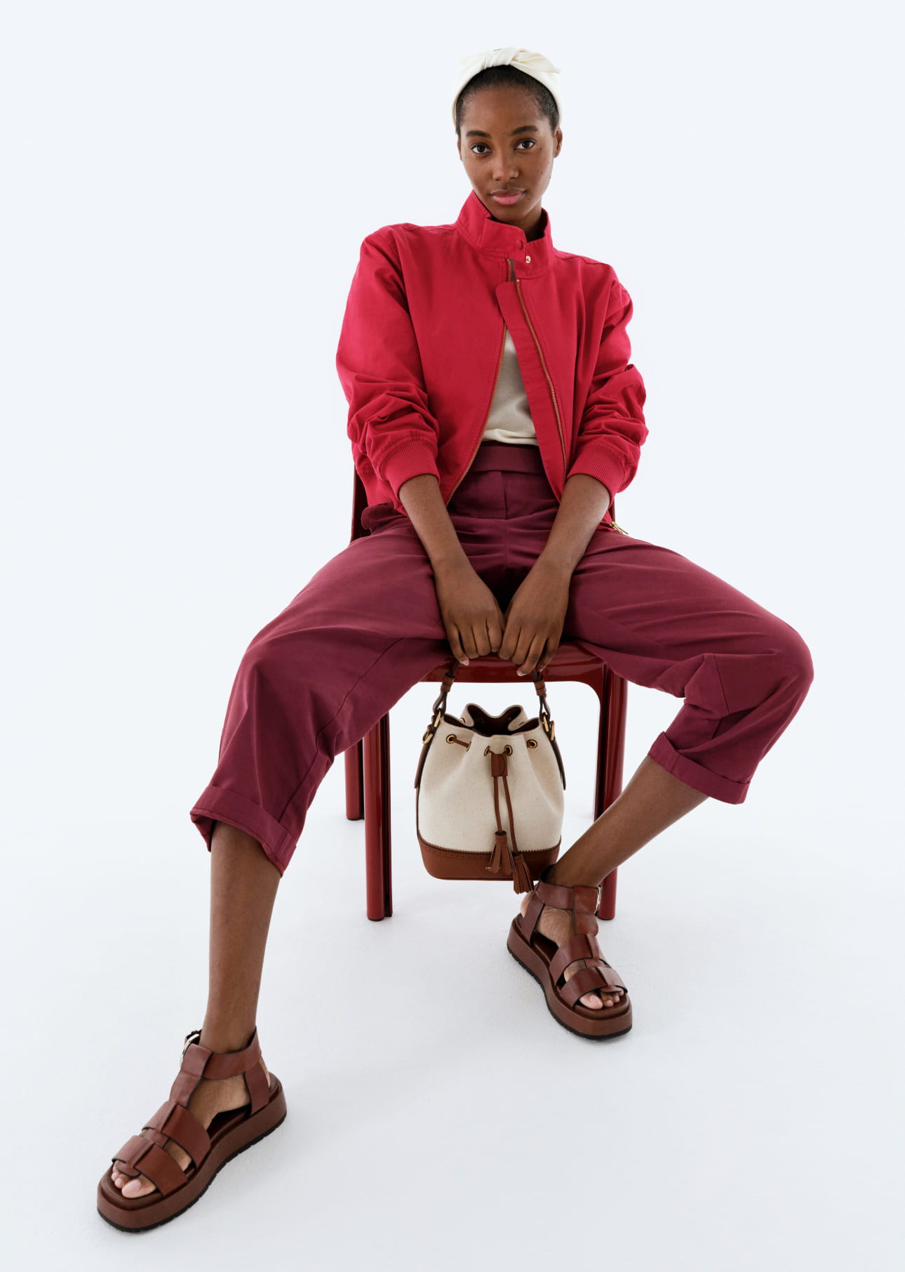 Model sitting on a chair, wearing red jacket and maroon pants, with sandals.
