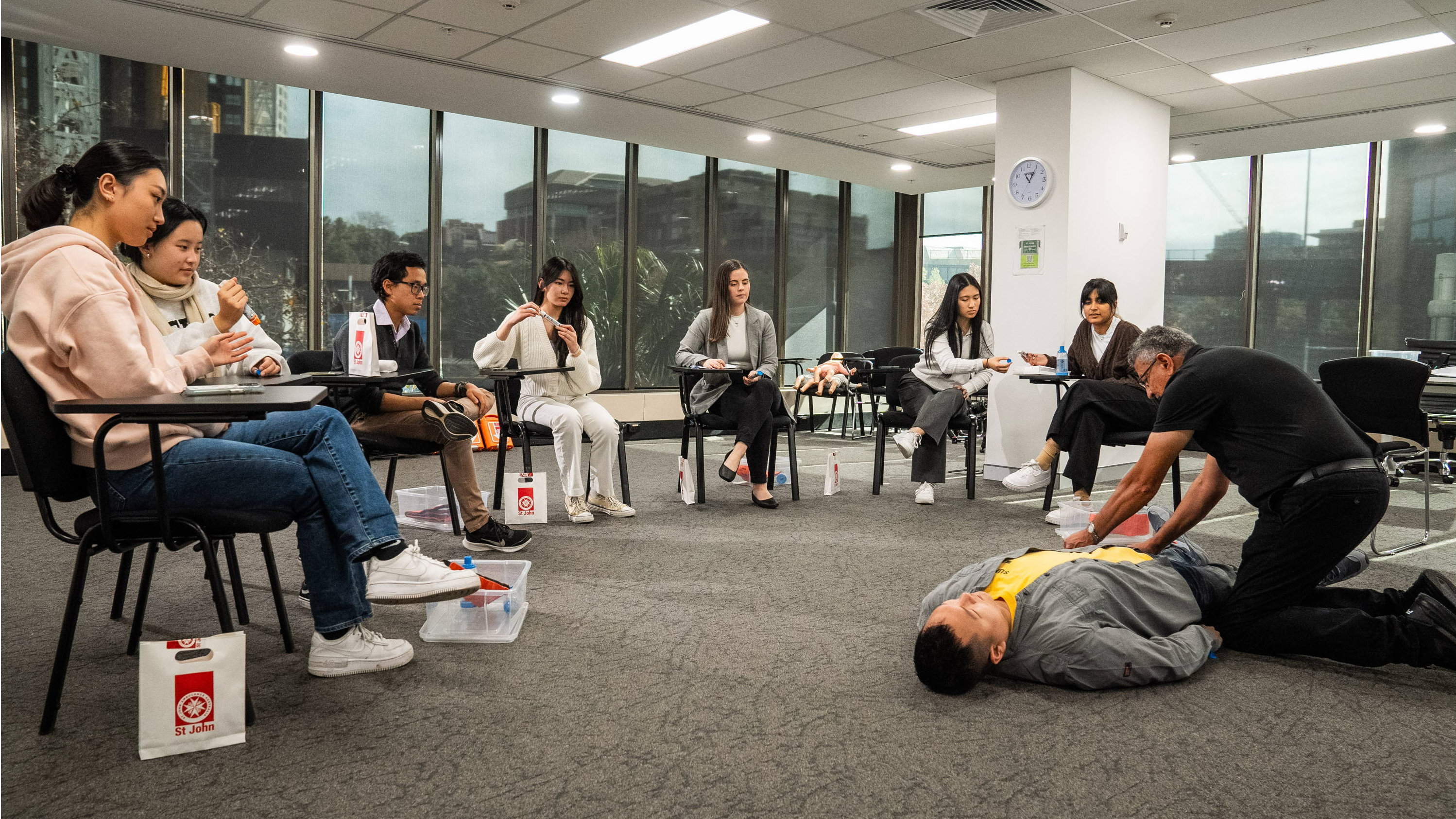 group of students sitting at individual tables watching a first aid trainer and volunteer demonstrate how to perform first aid.