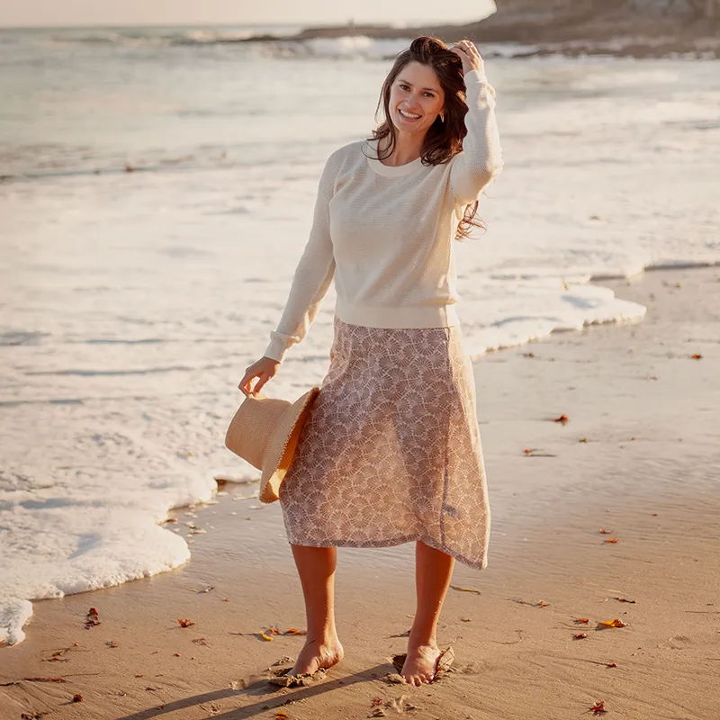 Woman in a sweater and skirt holding a hat on the beach.