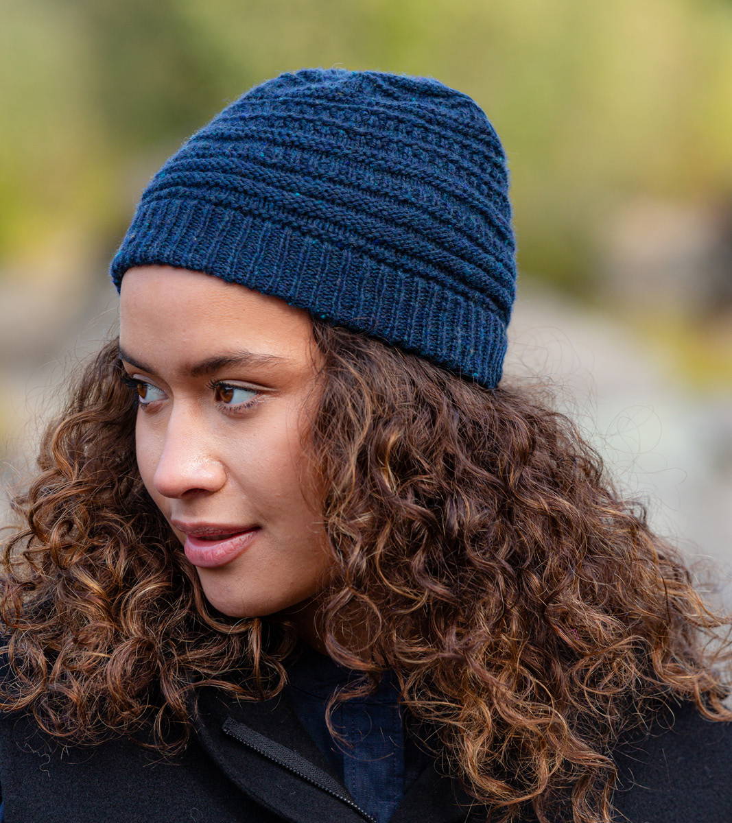Sadie, a woman with medium curly hair, models Brooklyn Tweed hand knit Grist Hat in Imbue Sport color Boro -- side view