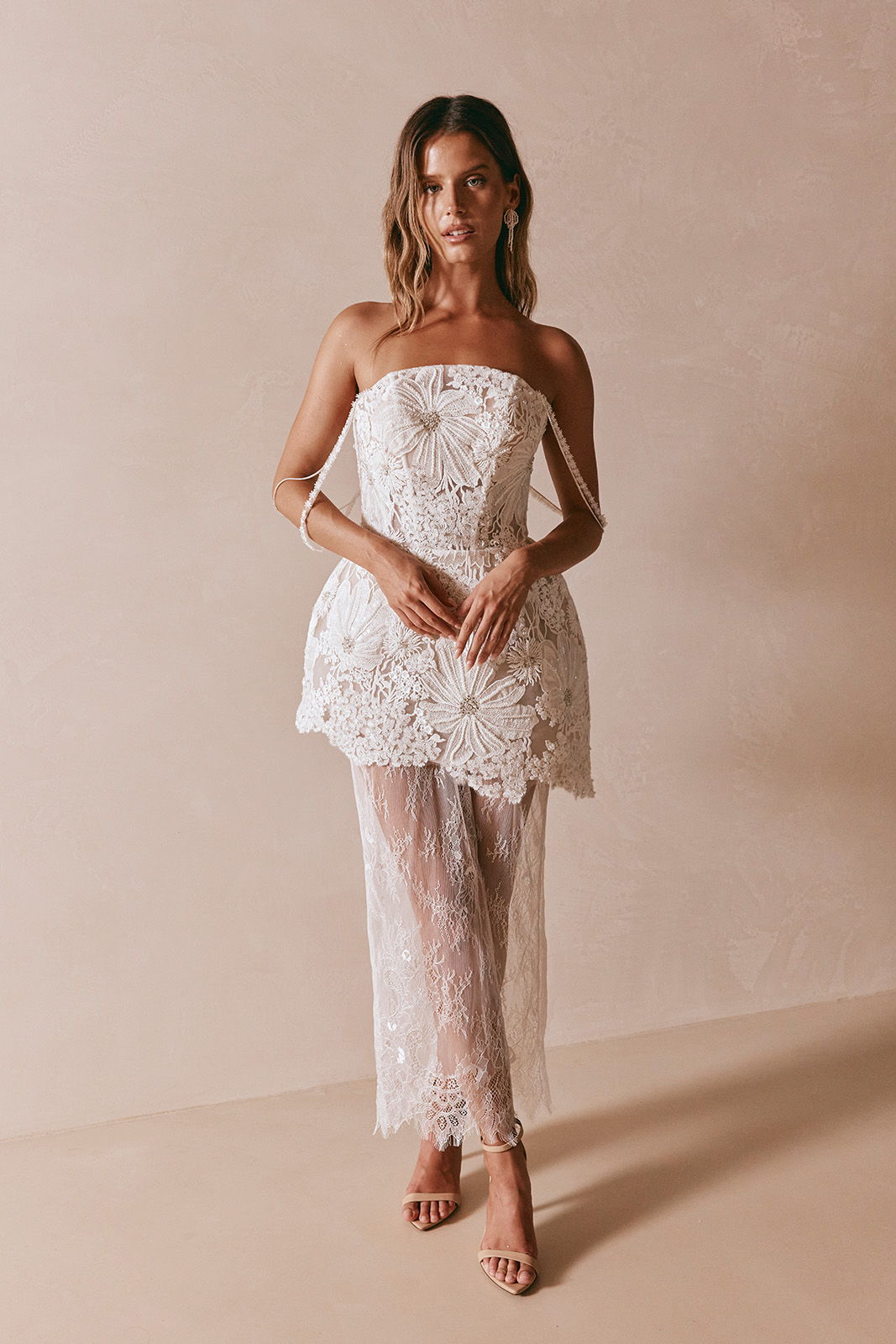 Woman in a strapless white lace dress standing against a neutral background.