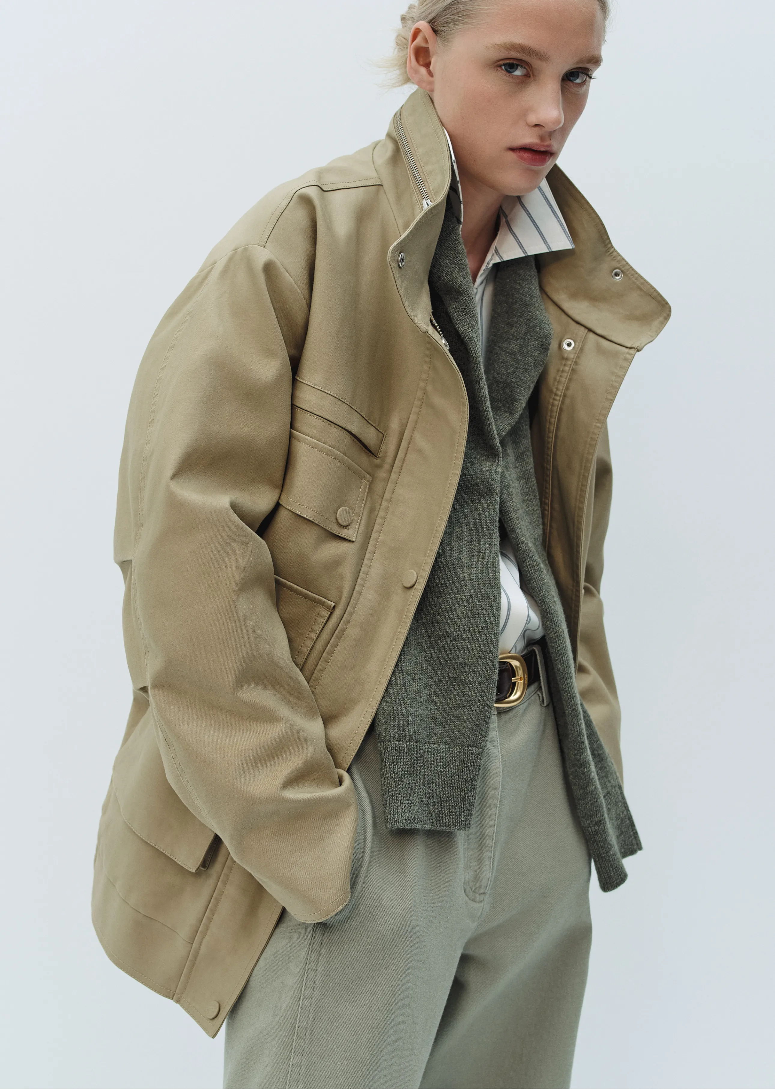 A model wearing a beige jacket over a green sweater and light pants.