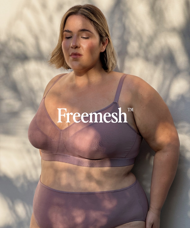 A plus-size model wearing a bra and underwear against a shadowed wall.