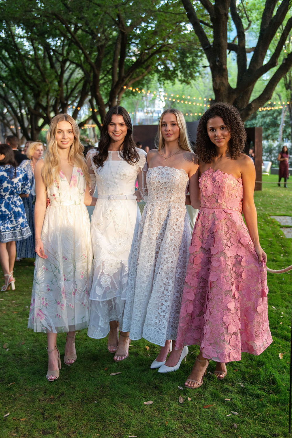 Four models in elegant dresses standing in a garden setting.
