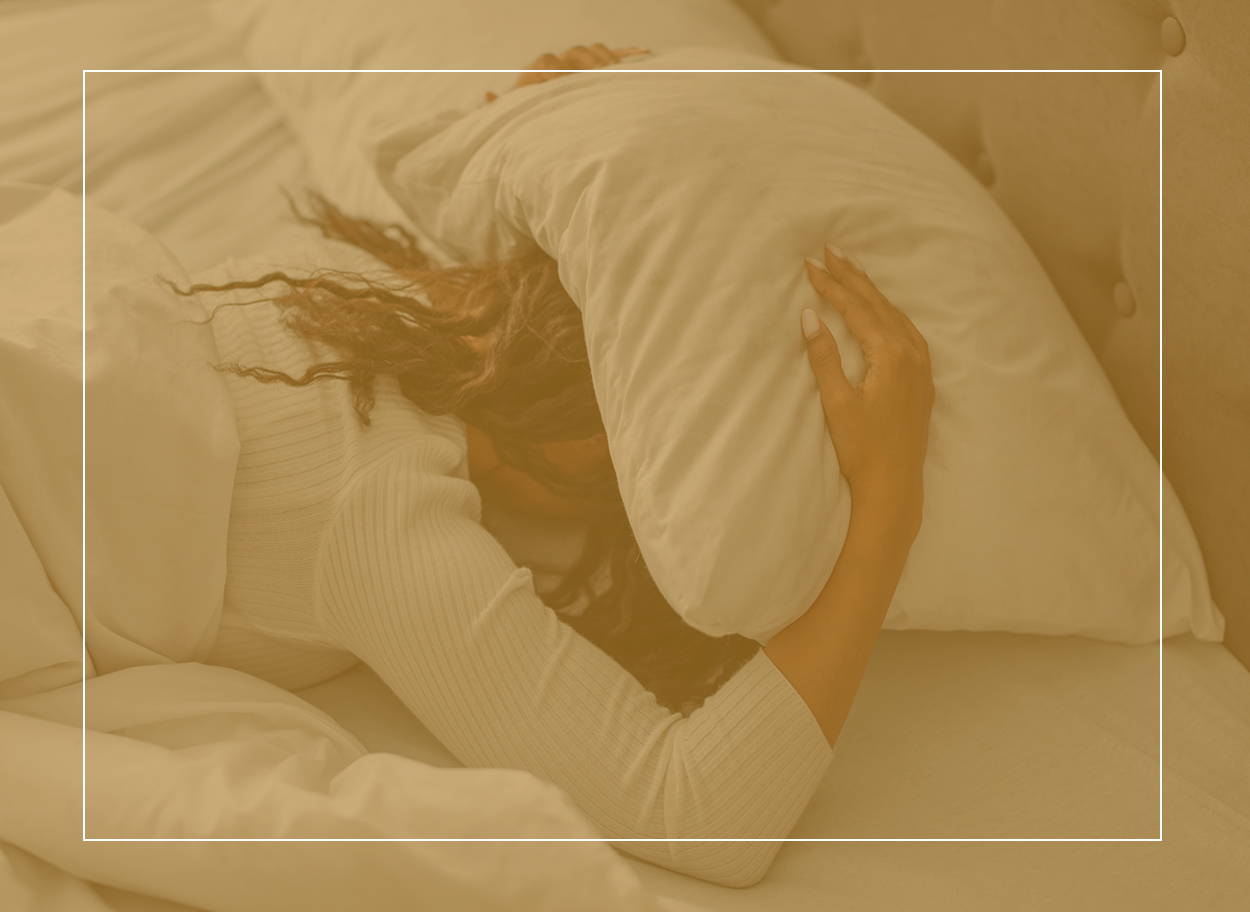 Eine Frau liegt nachts auf dem Bauch im Bett und hat ein Kissen über dem Kopf. Sie hat eine Hausstaubmilbenallergie und kann aufgrund von Symptomen nicht schlafen.