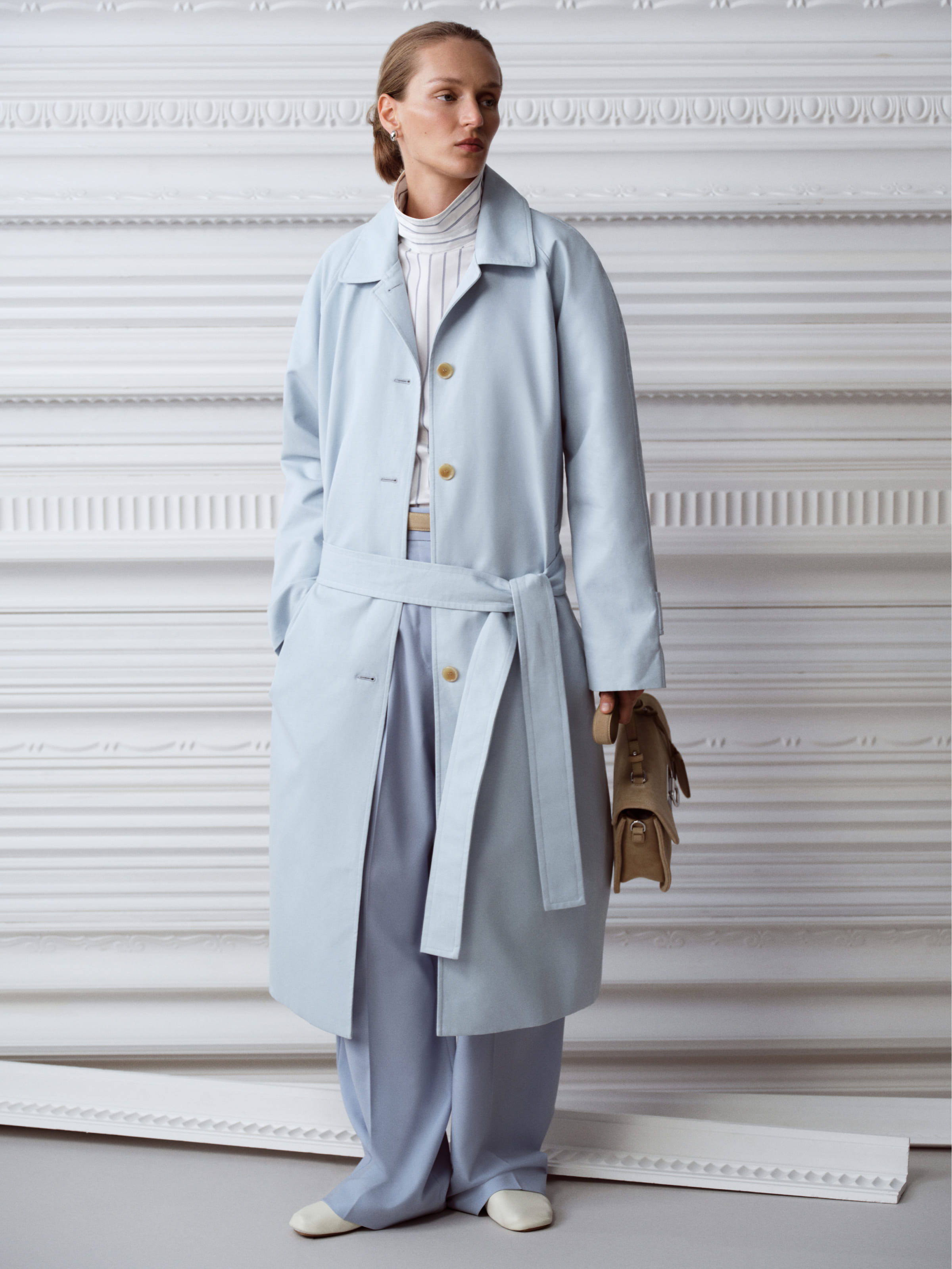 Fashion model wearing a light blue coat and trousers against a textured background.