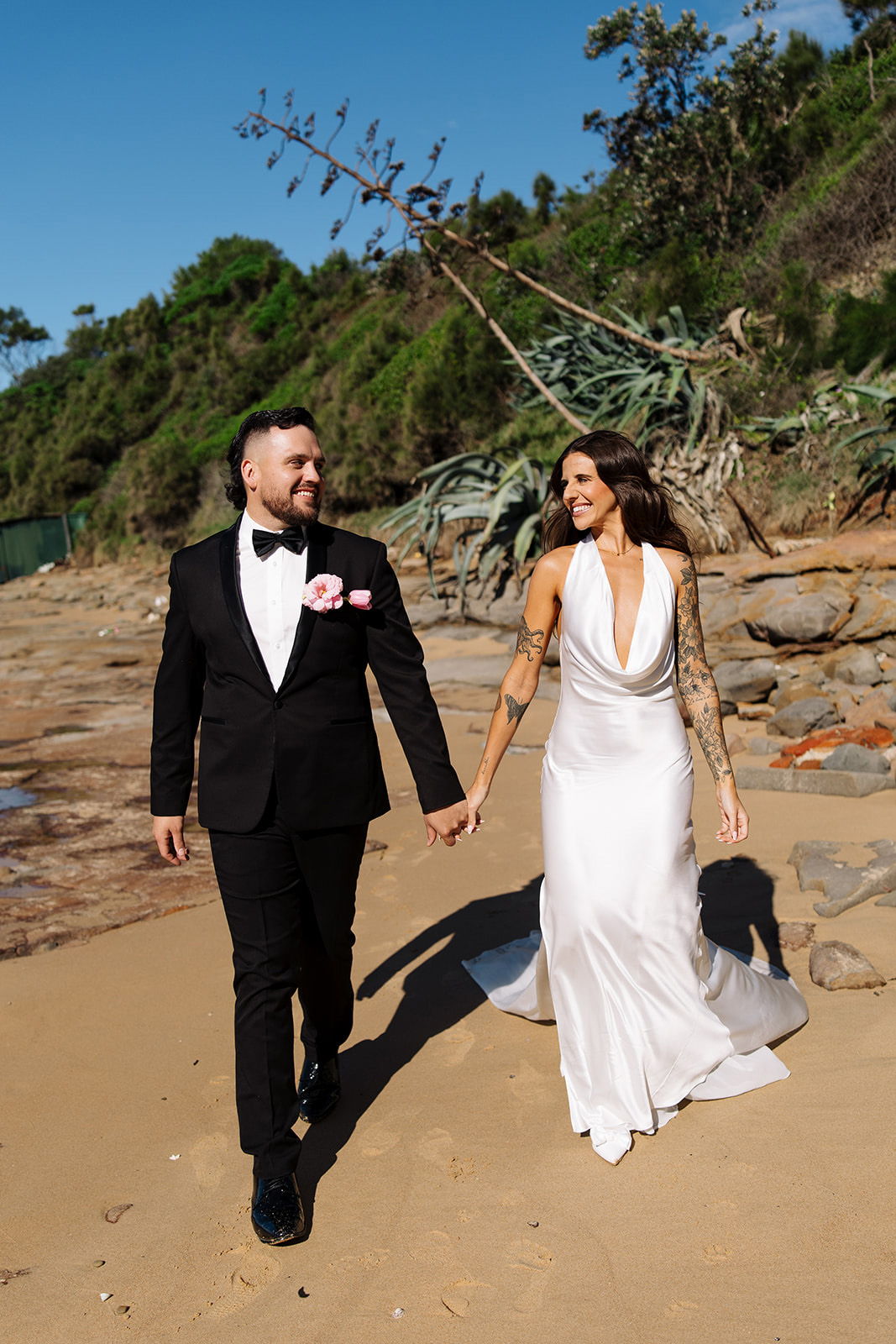 A couple walking hand in hand on the beach, wearing formal wedding attire.