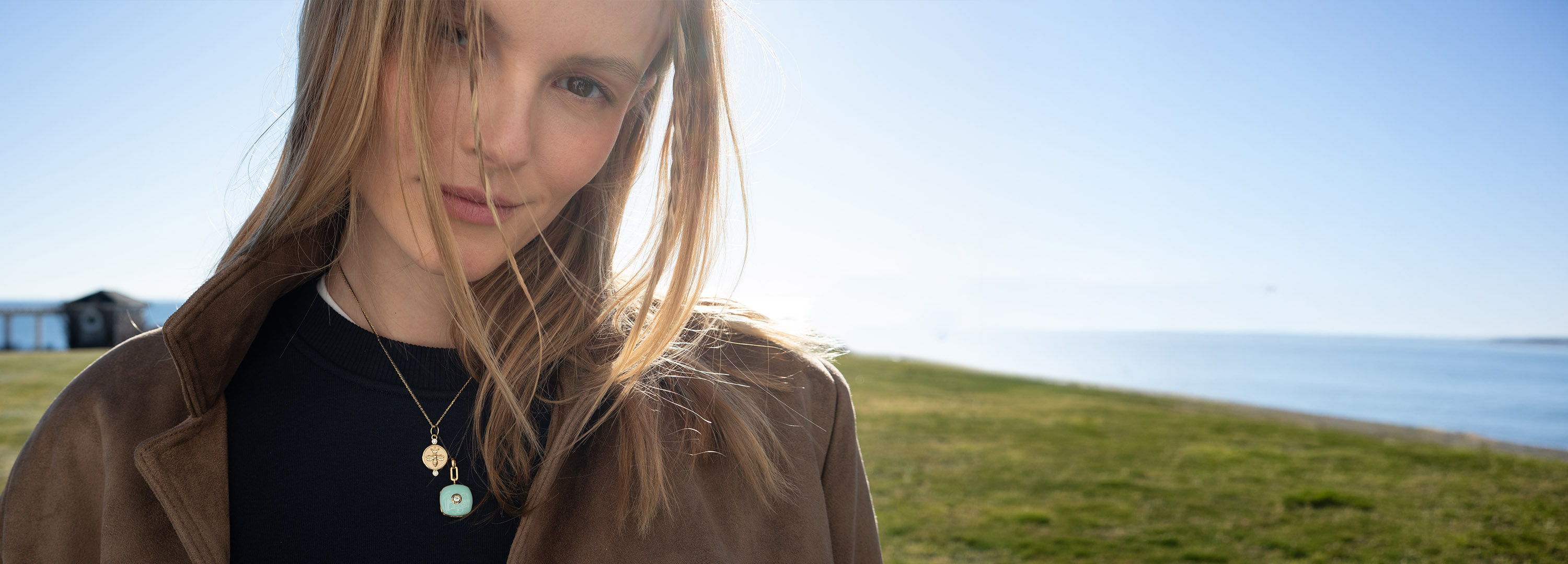 Woman with long hair, wearing a jacket, by the sea on a sunny day.