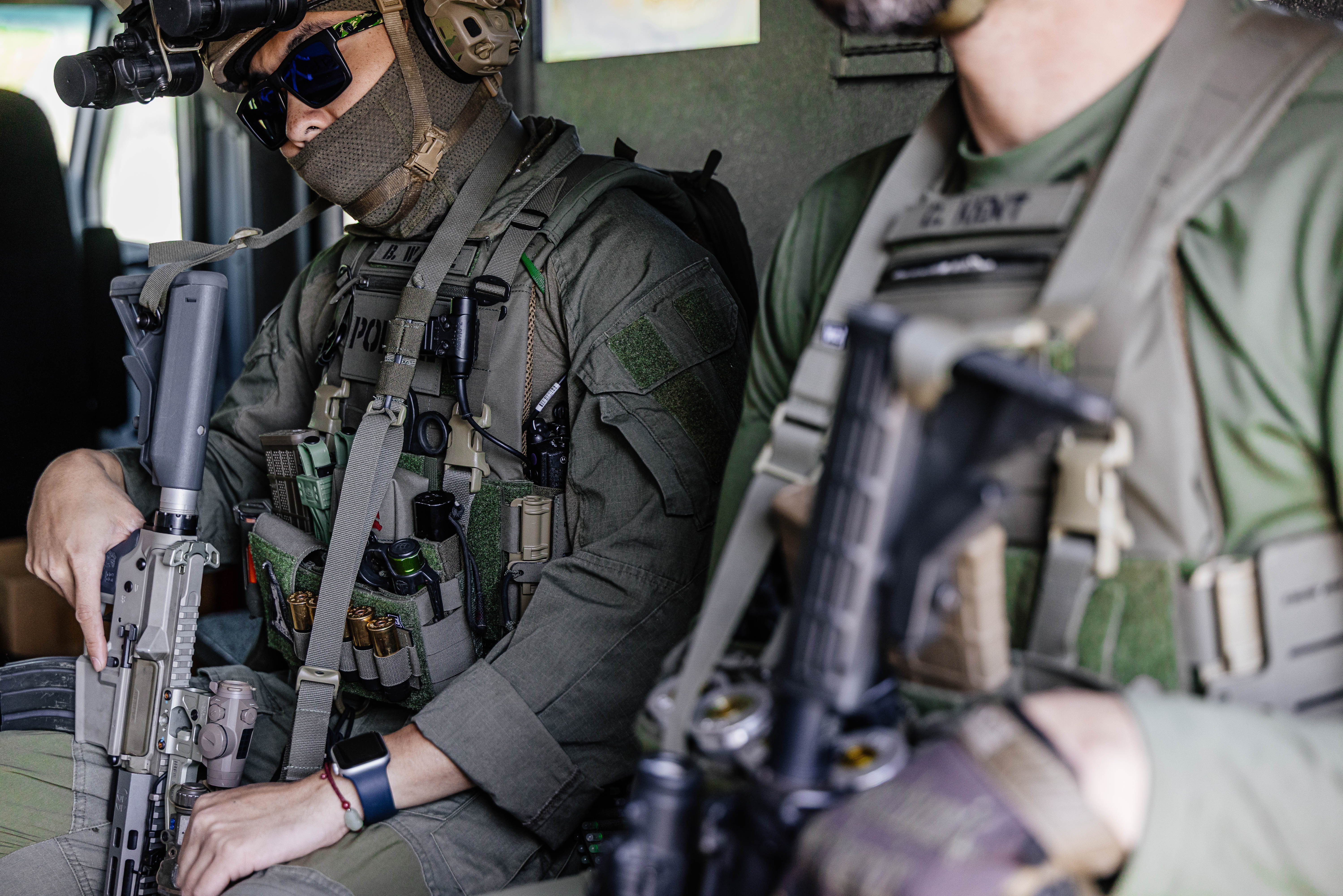 Two armed officers seated inside a tactical vehicle, wearing tactical gear.