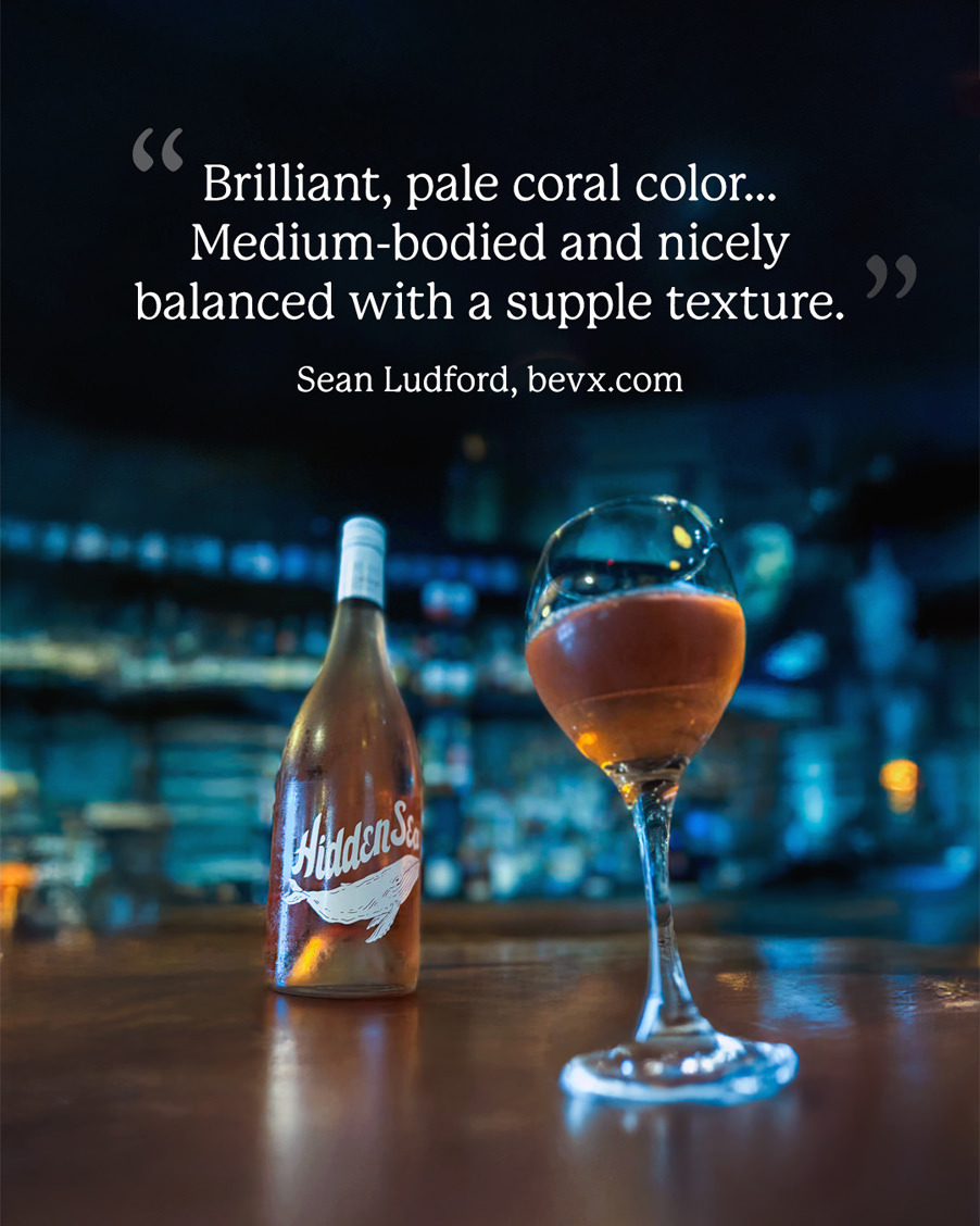 The Hidden Sea rosé bottle and a filled wine glass on a bar counter with a quote overlay: “Brilliant, pale coral color… Medium-bodied and nicely balanced with a supple texture.” — Sean Ludford, bevx.com.