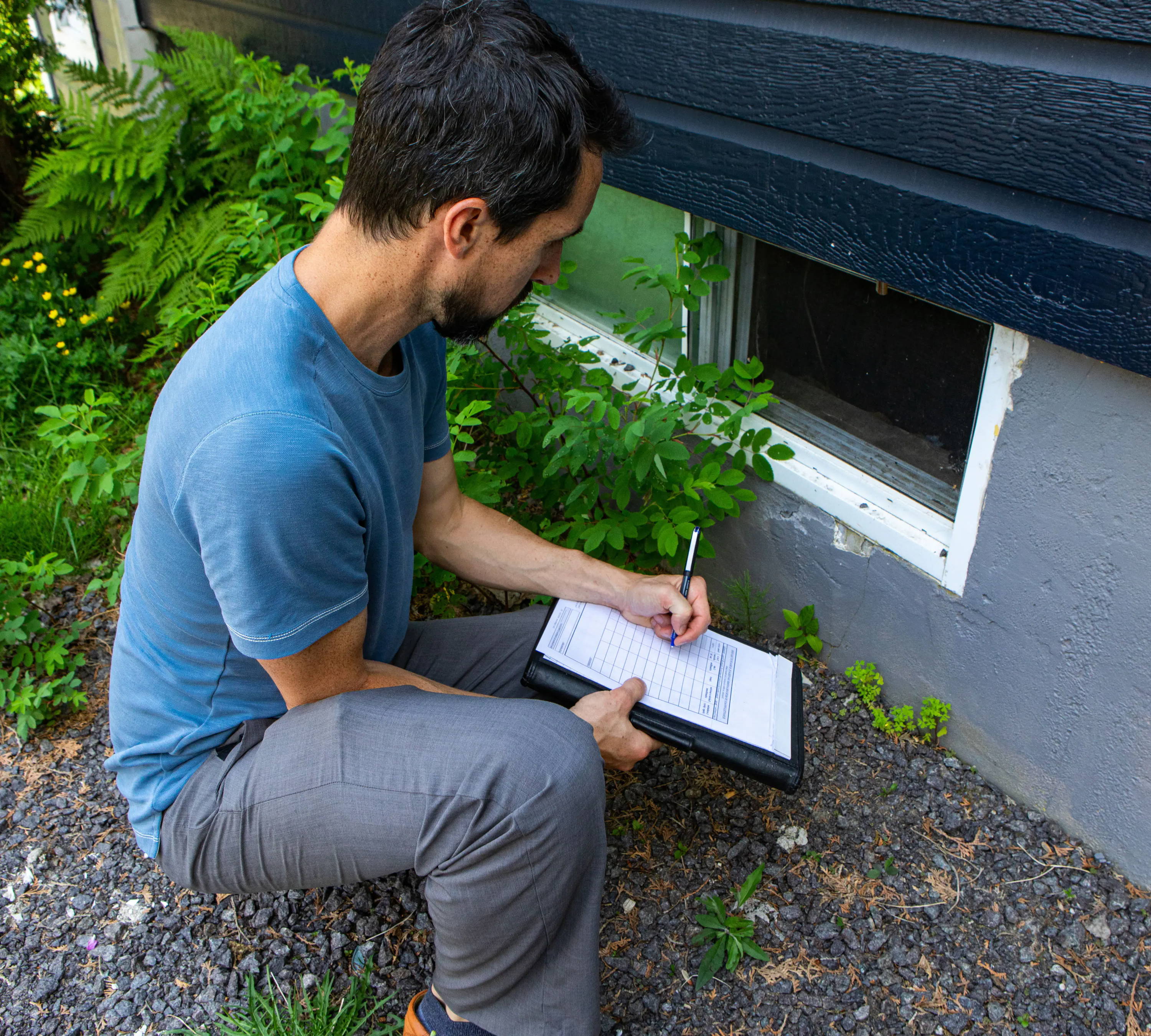 Person crouching by a house, taking notes near a window.