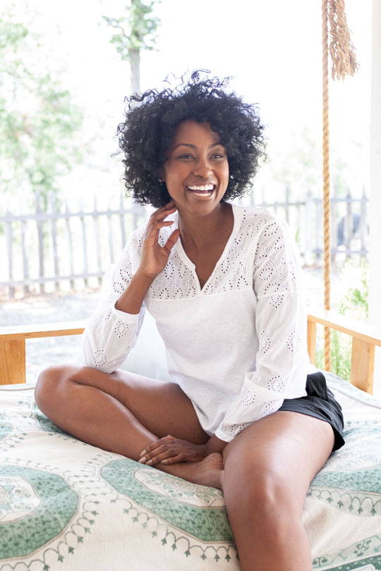 Woman sitting on swinging bed on porch wearing white Seychelle long sleeve top.