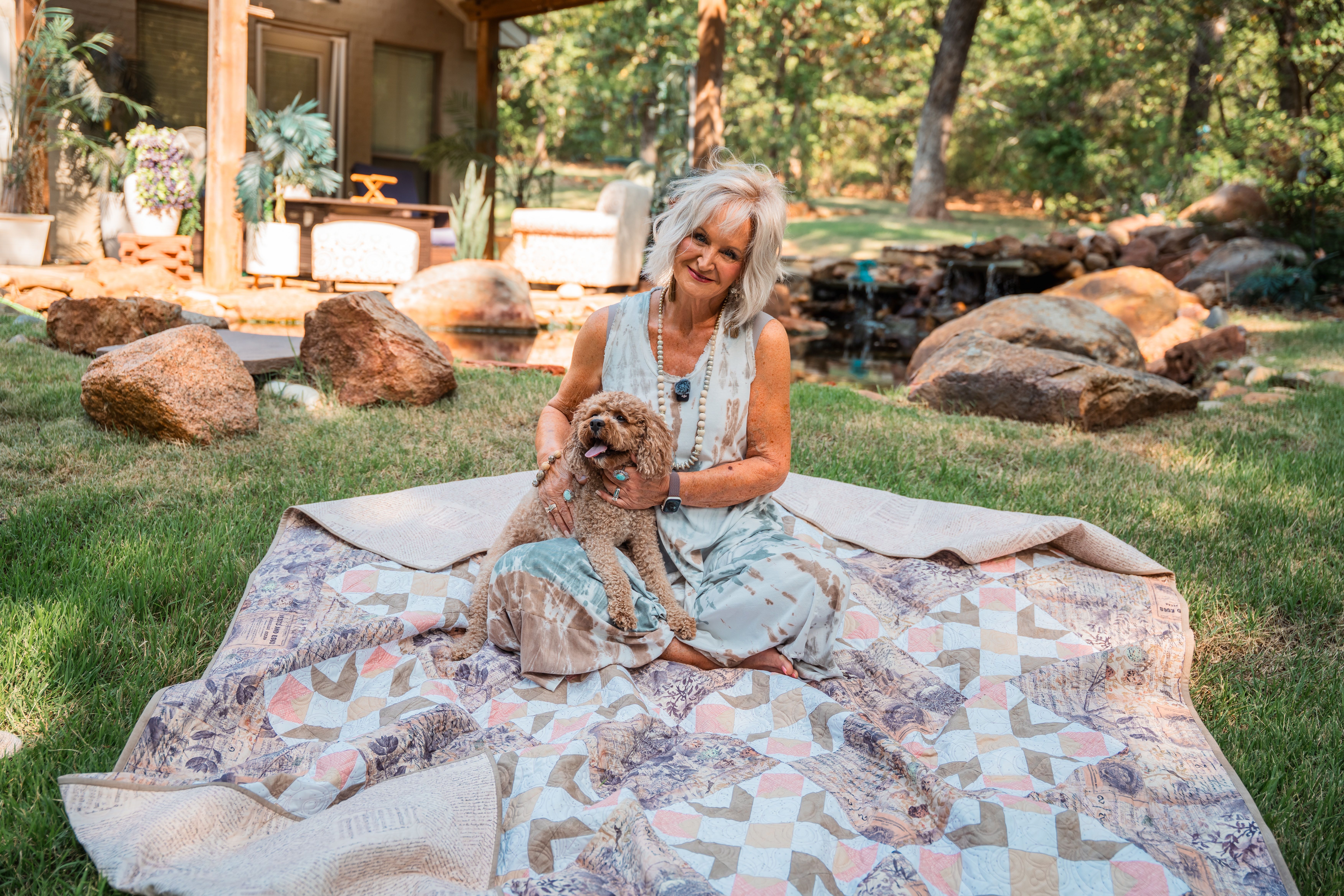 Woman sitting on a quilt with a dog in a garden setting.