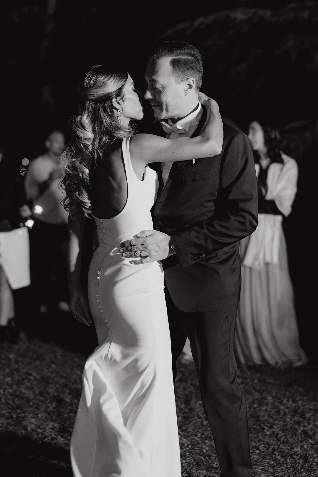 Couple dancing intimately at night, surrounded by guests in elegant attire.