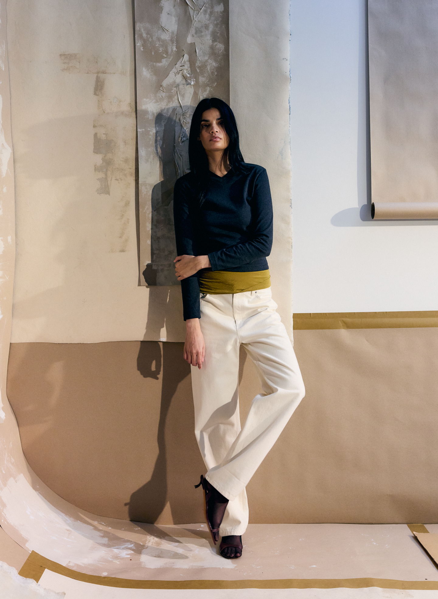 Model wearing a dark top and white pants, leaning against a wall.